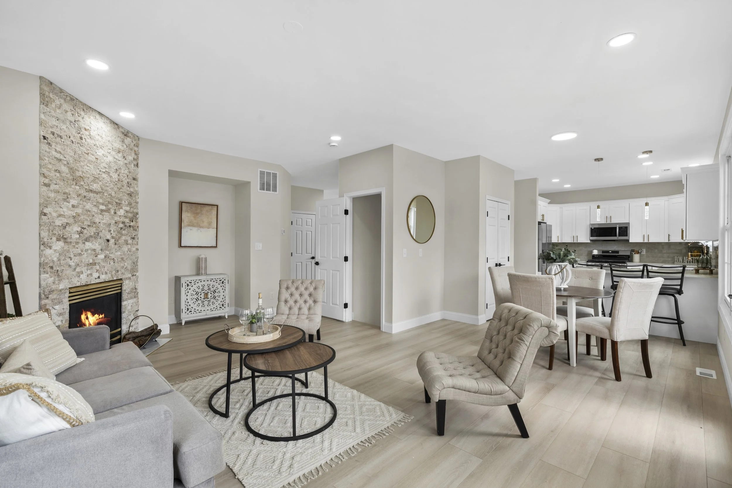 Open-concept living room and kitchen with beige walls, white cabinetry, a brick fireplace, and modern furniture including a gray sofa, beige armchairs, and black coffee tables.