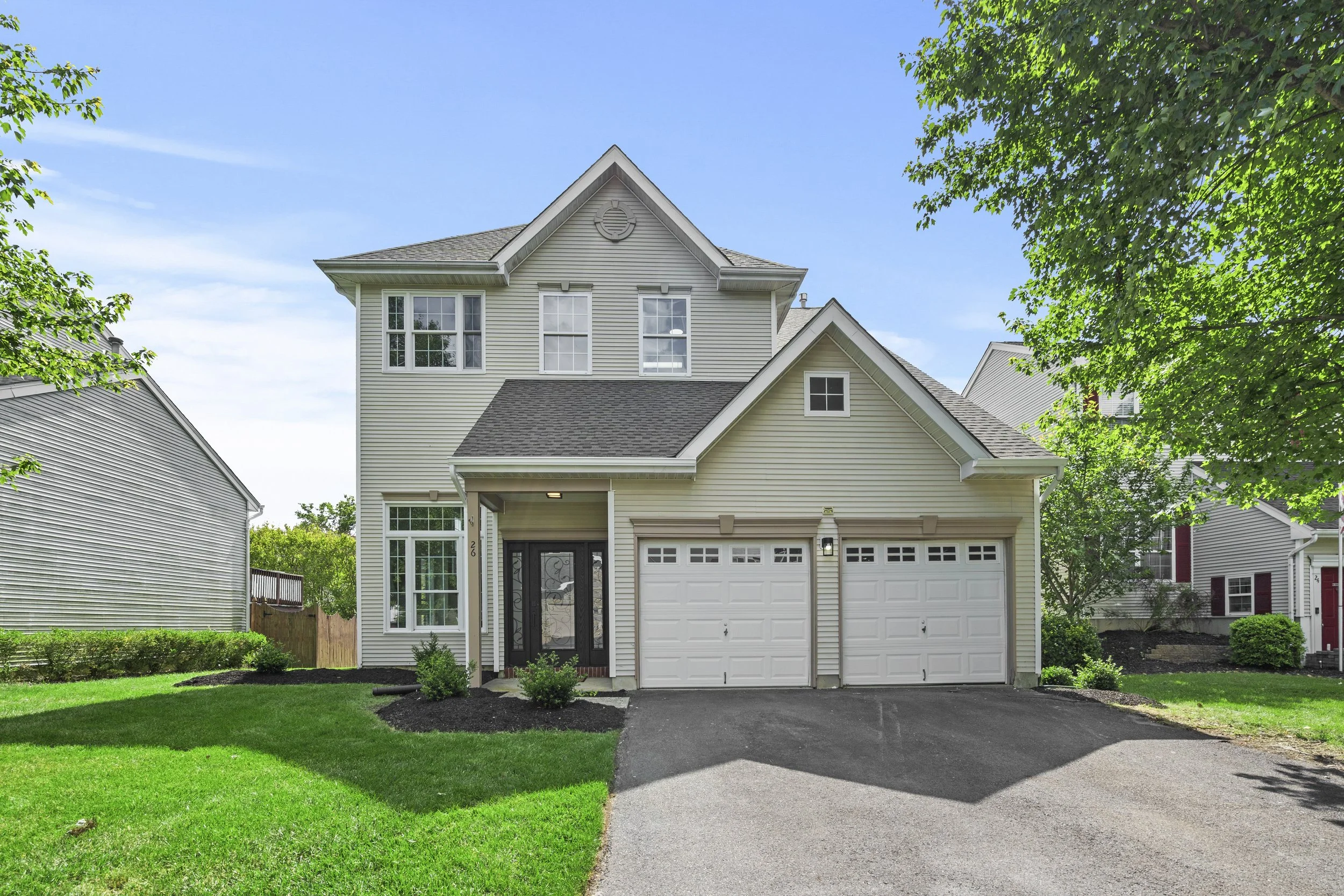 A modern two-story house with beige siding, a gray shingled roof, and white trim. It has a black front door with glass panels and a white garage door. The front yard has a driveway, green grass, and small bushes. There are trees with green leaves and