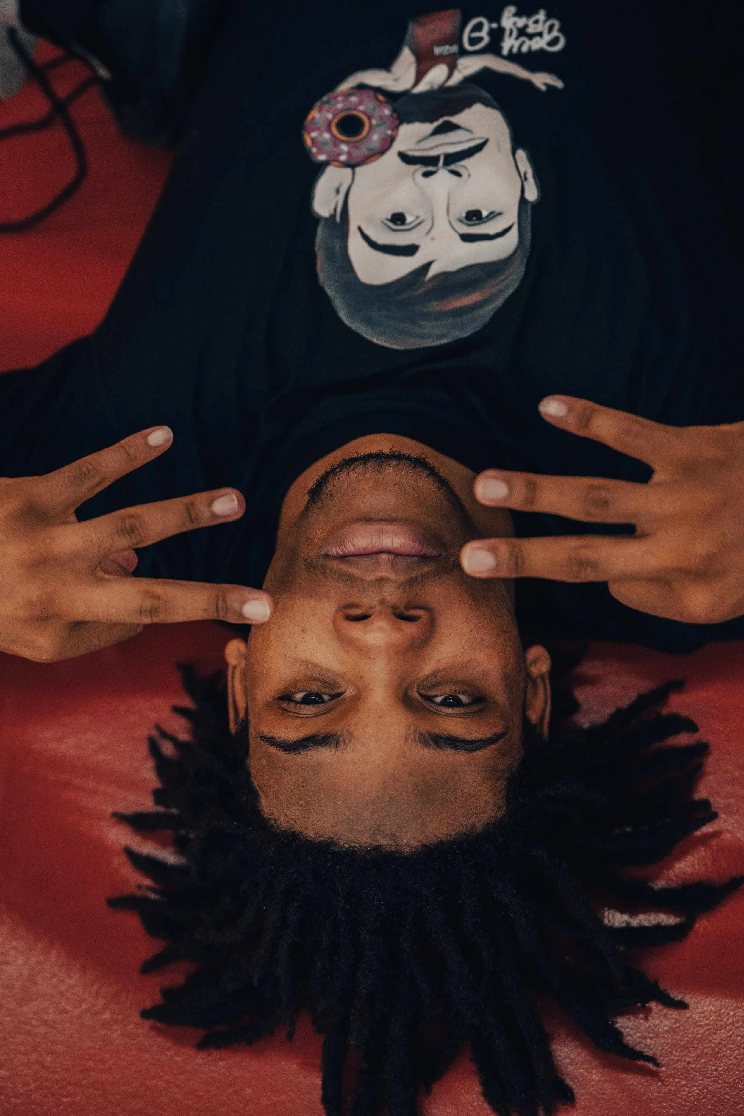 A young person with curly black hair lying on their back on a red surface, making a peace sign with their fingers on both hands, and holding a painted face mask on their chest depicting a woman with dark hair and a donut on her face.