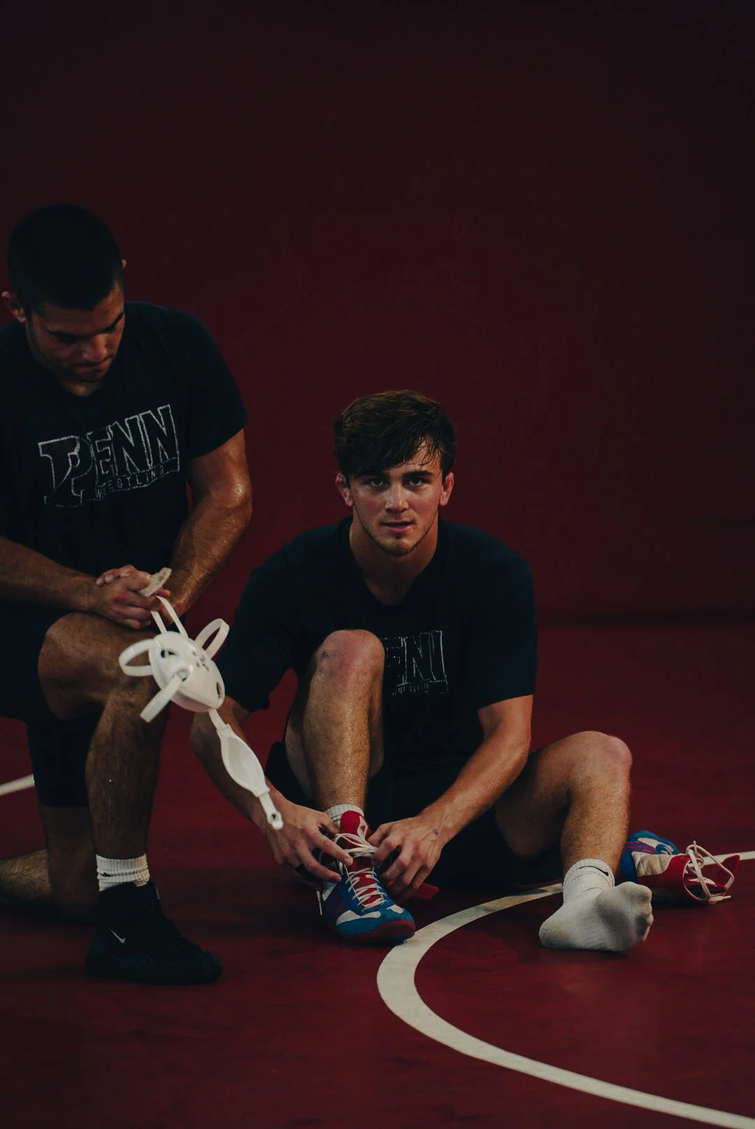 A young male wrestler sitting on a maroon wrestling mat, with another male assistant tying his shoelaces, both wearing black t-shirts with Penn Wrestling logo, indoor sports facility environment.