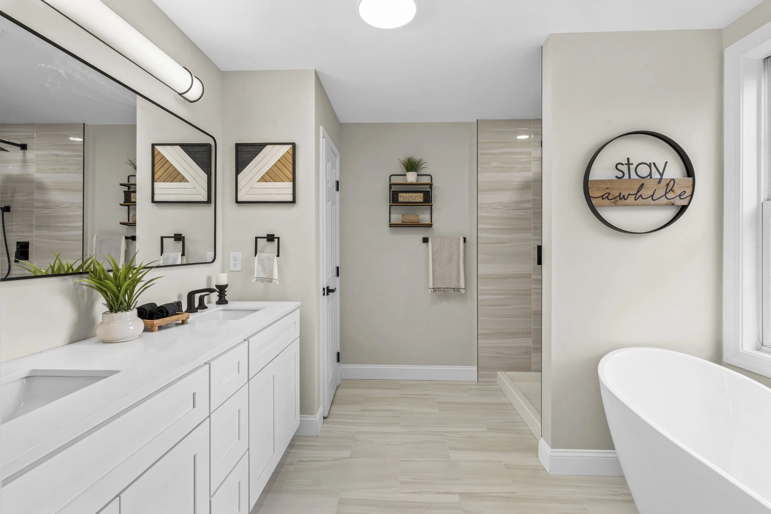 A modern bathroom with a white vanity and black fixtures, framed mirrors, beige wall tiles, a decorative plant on the countertop, a wall shelf with a potted plant and a sign that says 'Thankful,' a towel rack with a beige towel, a round wall sign tha