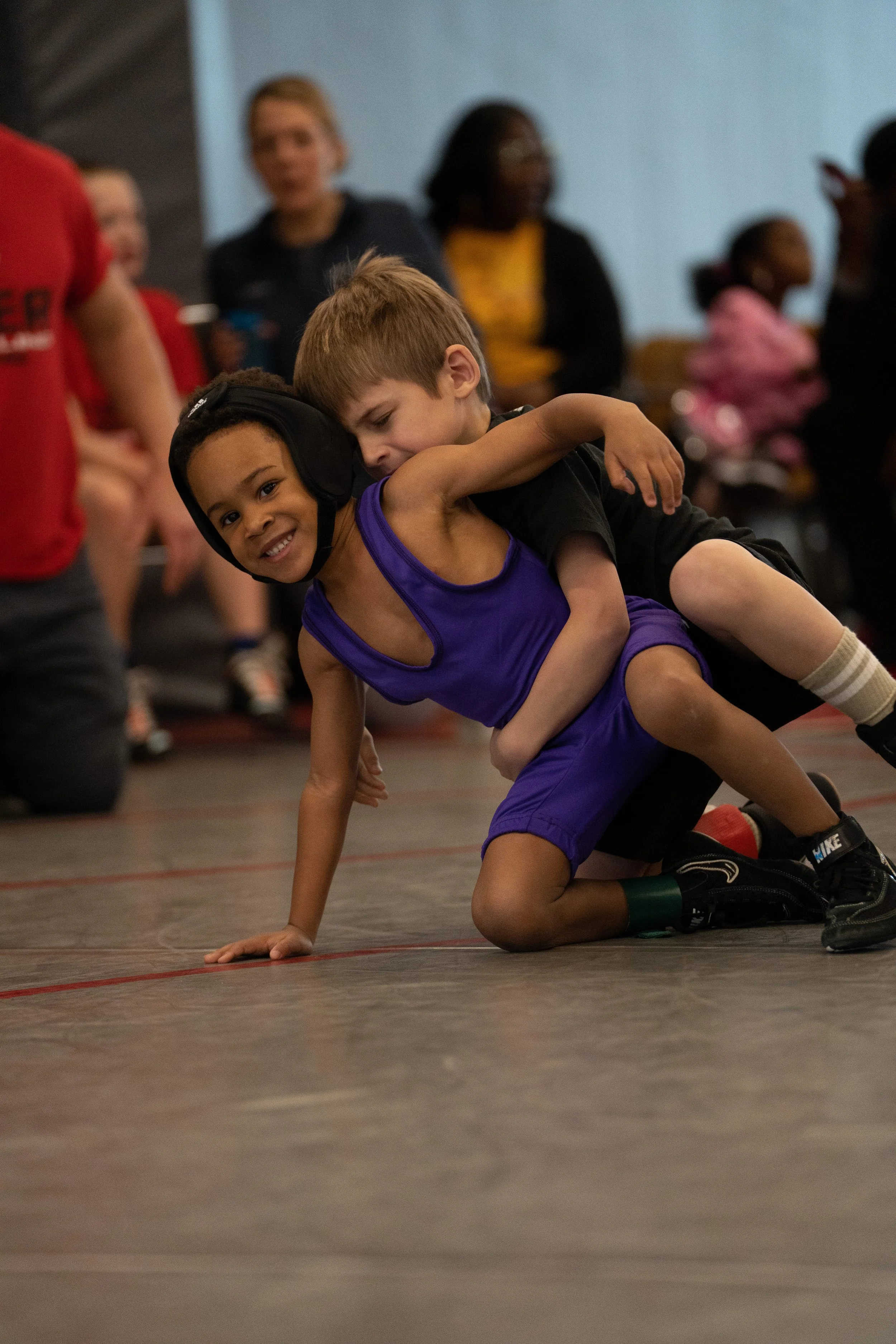 Two young boys wrestling on a gym floor, with other children and adults in the background.