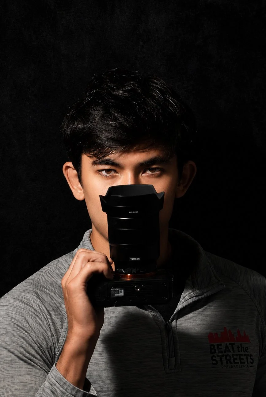 Young man taking a selfie with a camera and lens, wearing a gray zip-up jacket with 'BEAT THE STREETS PHILADELPHIA' logo, against a black background.