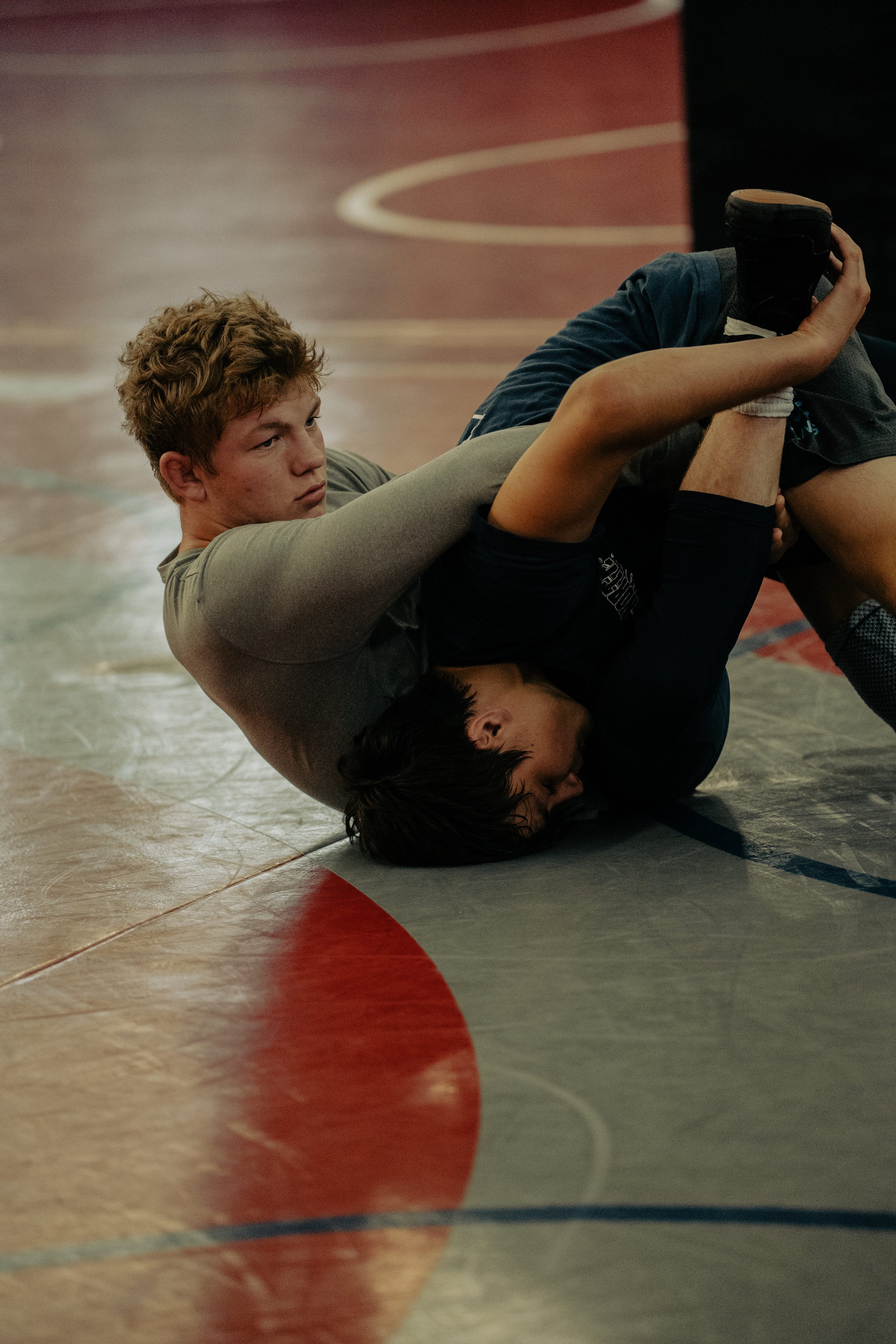 Two young men are practicing Brazilian Jiu-Jitsu on a gym mat, with one on top of the other and both engaged in a grappling position, in a martial arts training session.