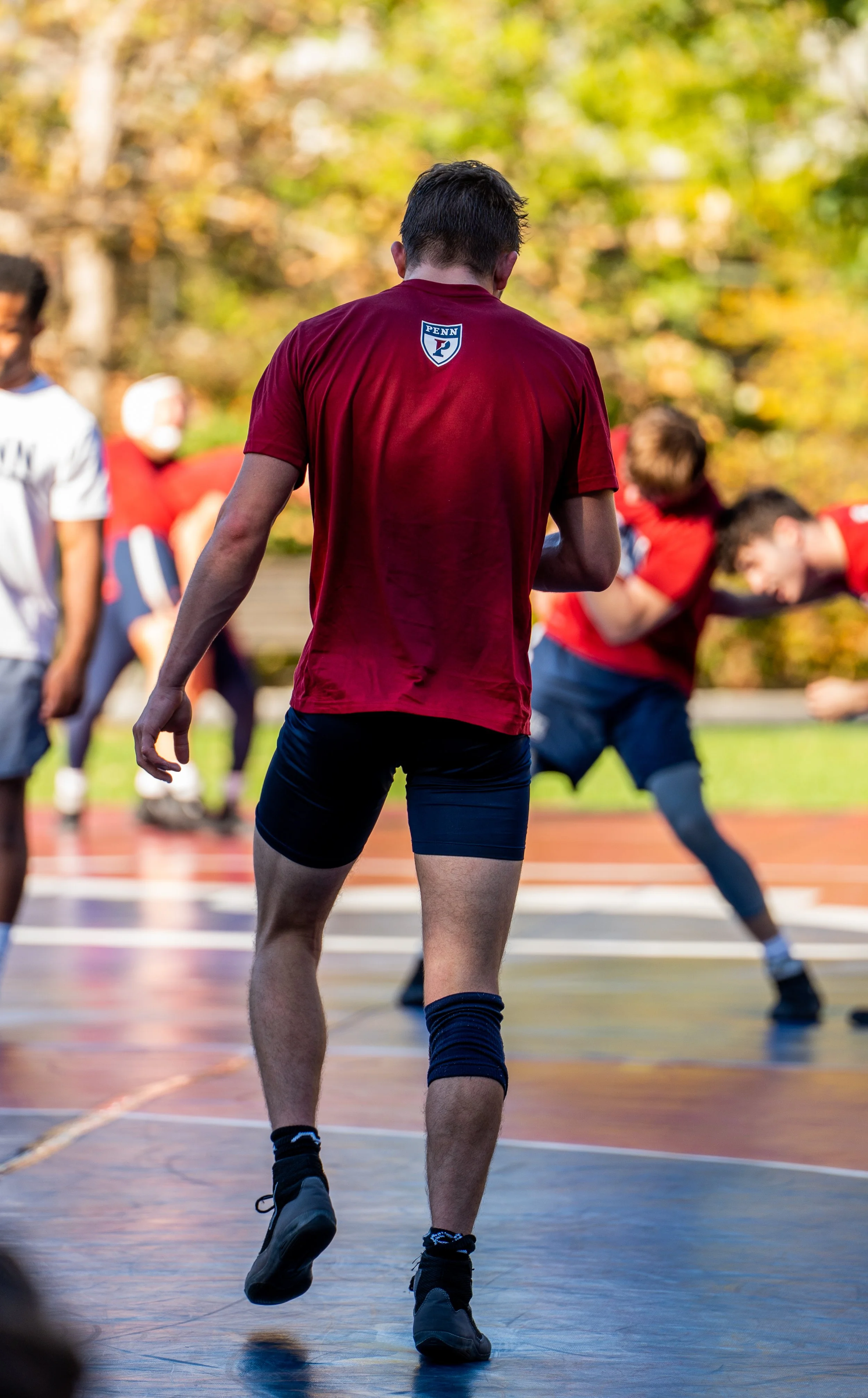 A basketball player from behind, wearing a red Penn shirt, on an outdoor court with other players in the background during an autumn day.