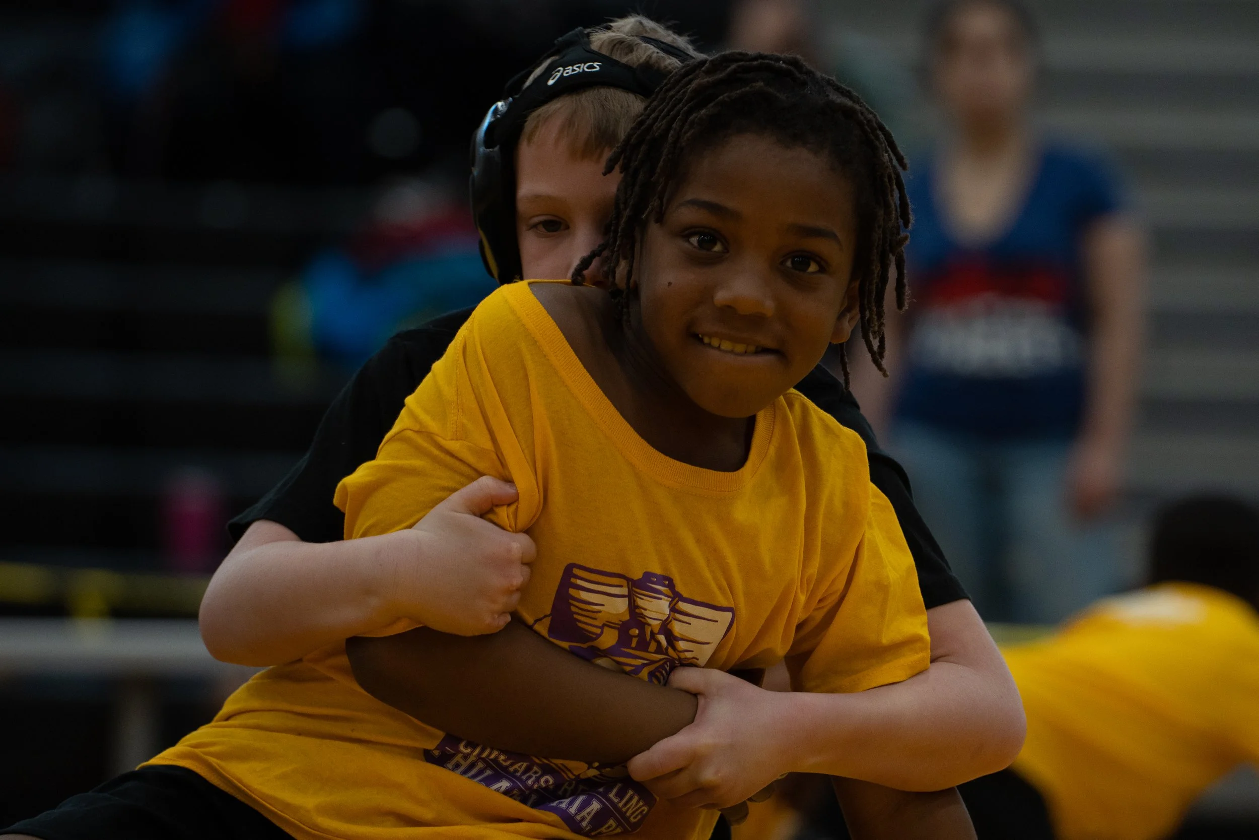 Two boys, one with dreadlocks and one with short light-colored hair wearing a black headband, embrace during a wrestling match in a gym.