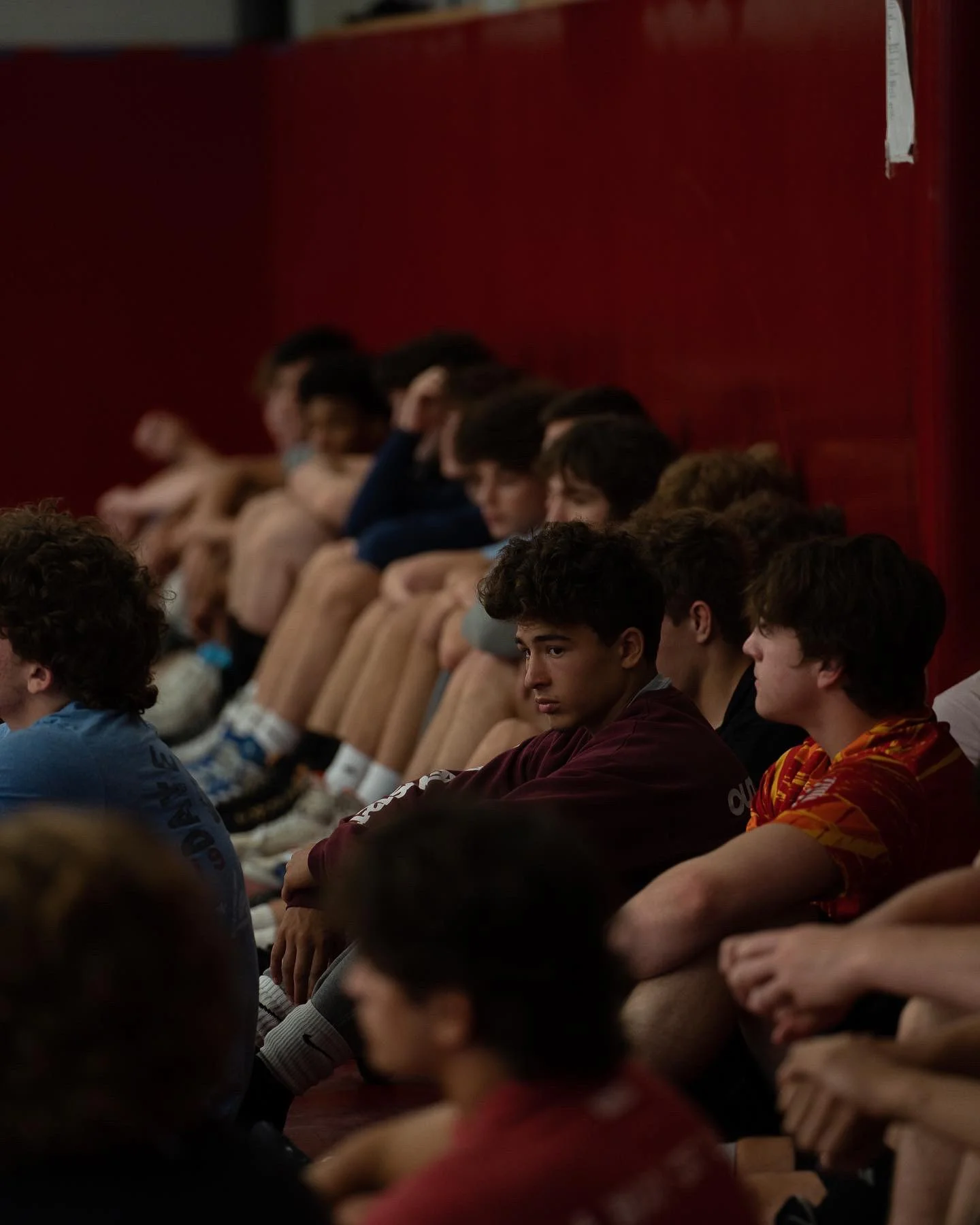 A group of young people sitting in auditorium chairs against a dark red wall, some appearing to be listening attentively, others looking engaged or distracted.