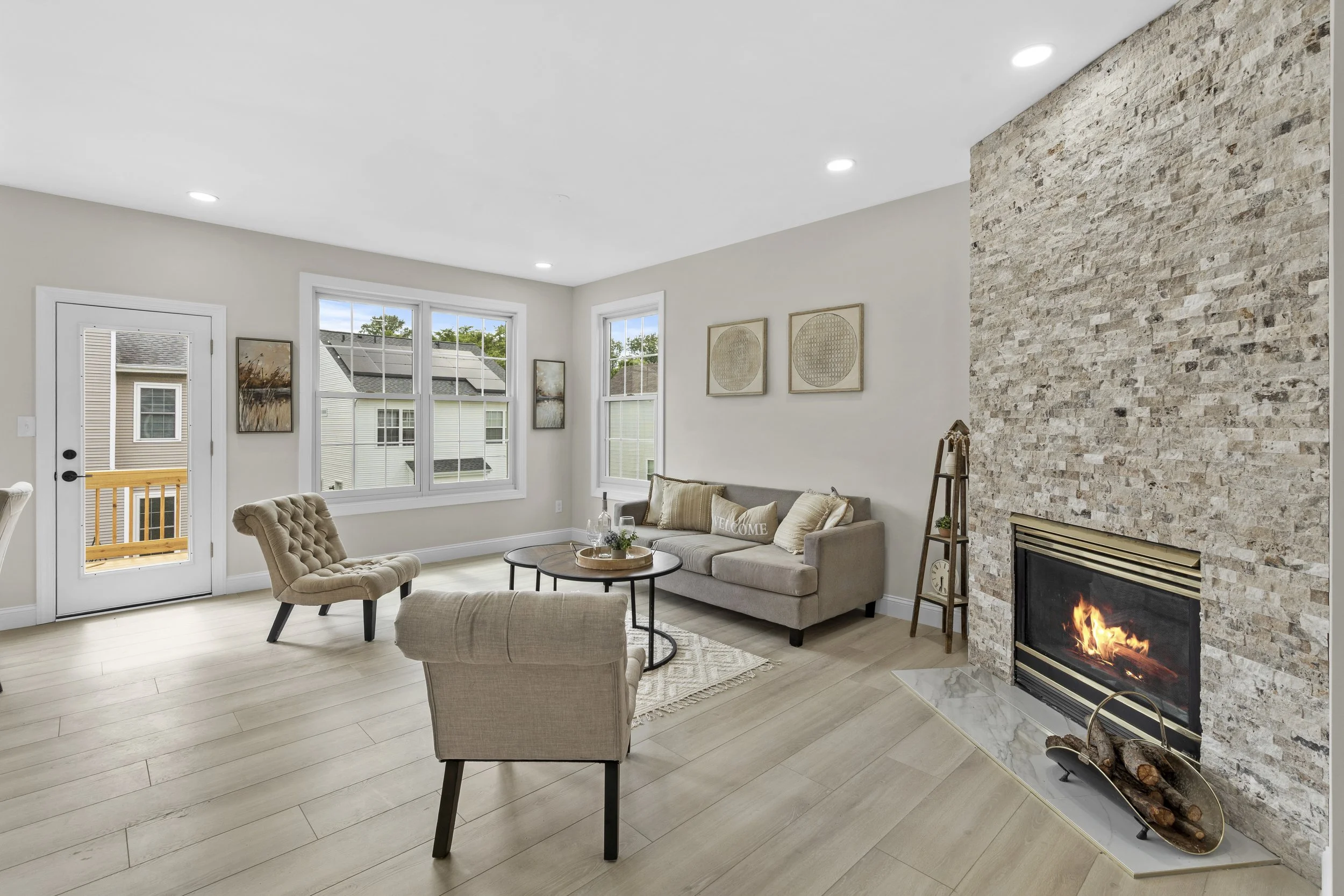 Living room with beige furniture, large windows, a brick fireplace with a fire burning, wall art, and a door leading to an outdoor balcony.