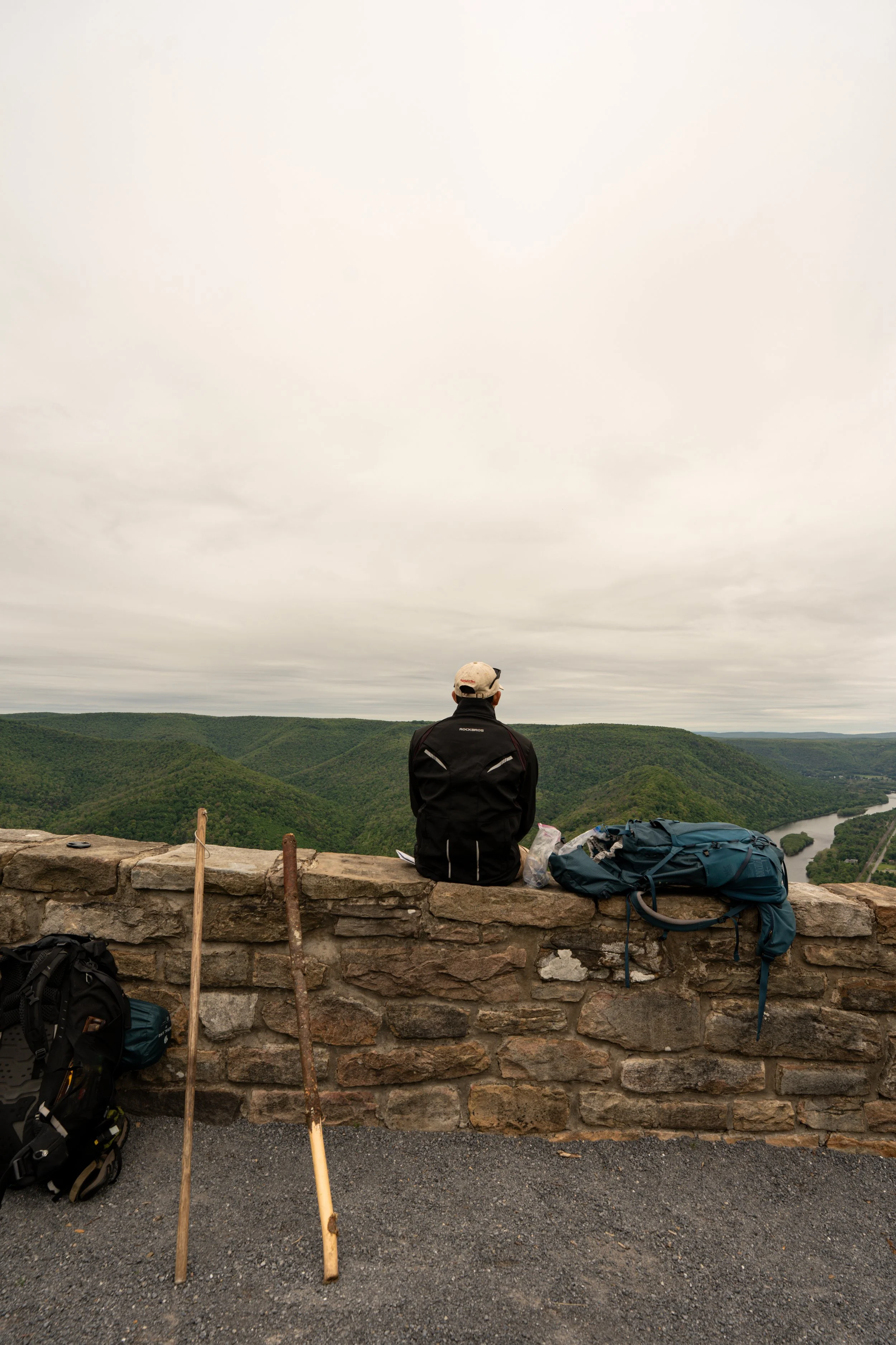 A person sitting on a stone wall overlooking green rolling hills and a river, wearing a hat and black jacket, with backpacks and hiking poles nearby.
