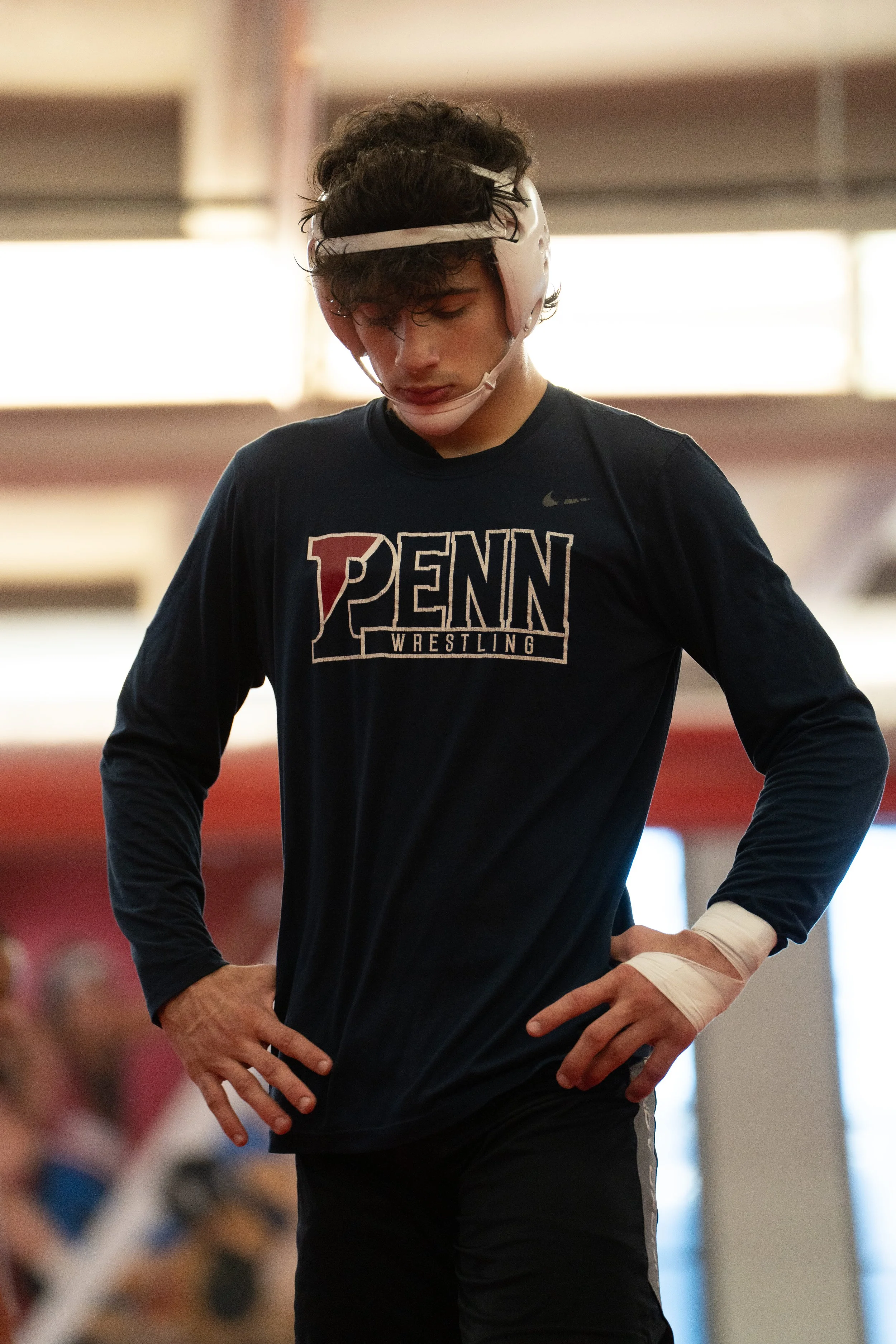 A male wrestler wearing a PENN Wrestling shirt and protective headgear, with his hands on his hips, inside a gymnasium.