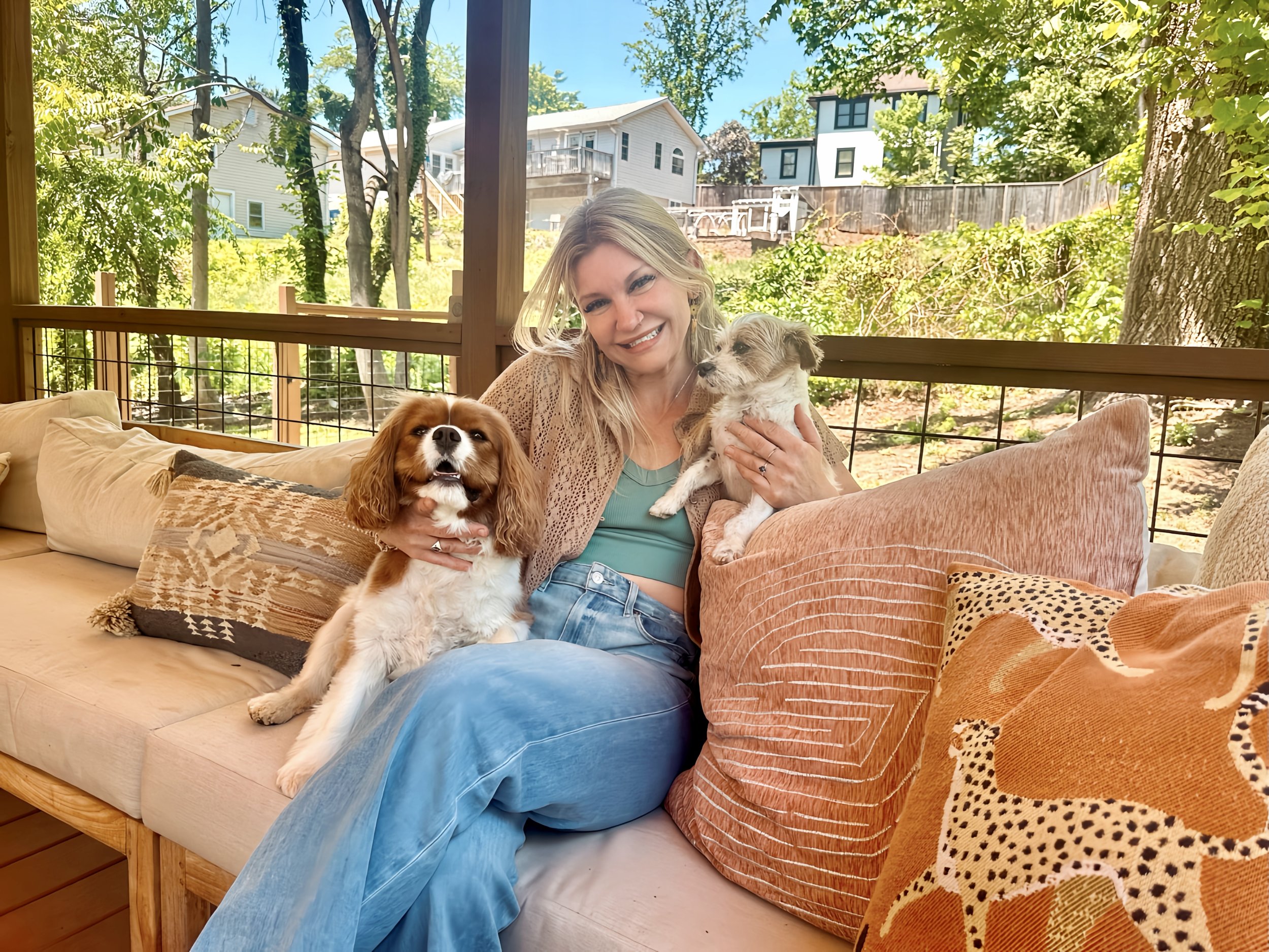 A woman sitting on a couch outdoors, holding two small dogs, smiling. Behind her are trees and houses in a suburban neighborhood.