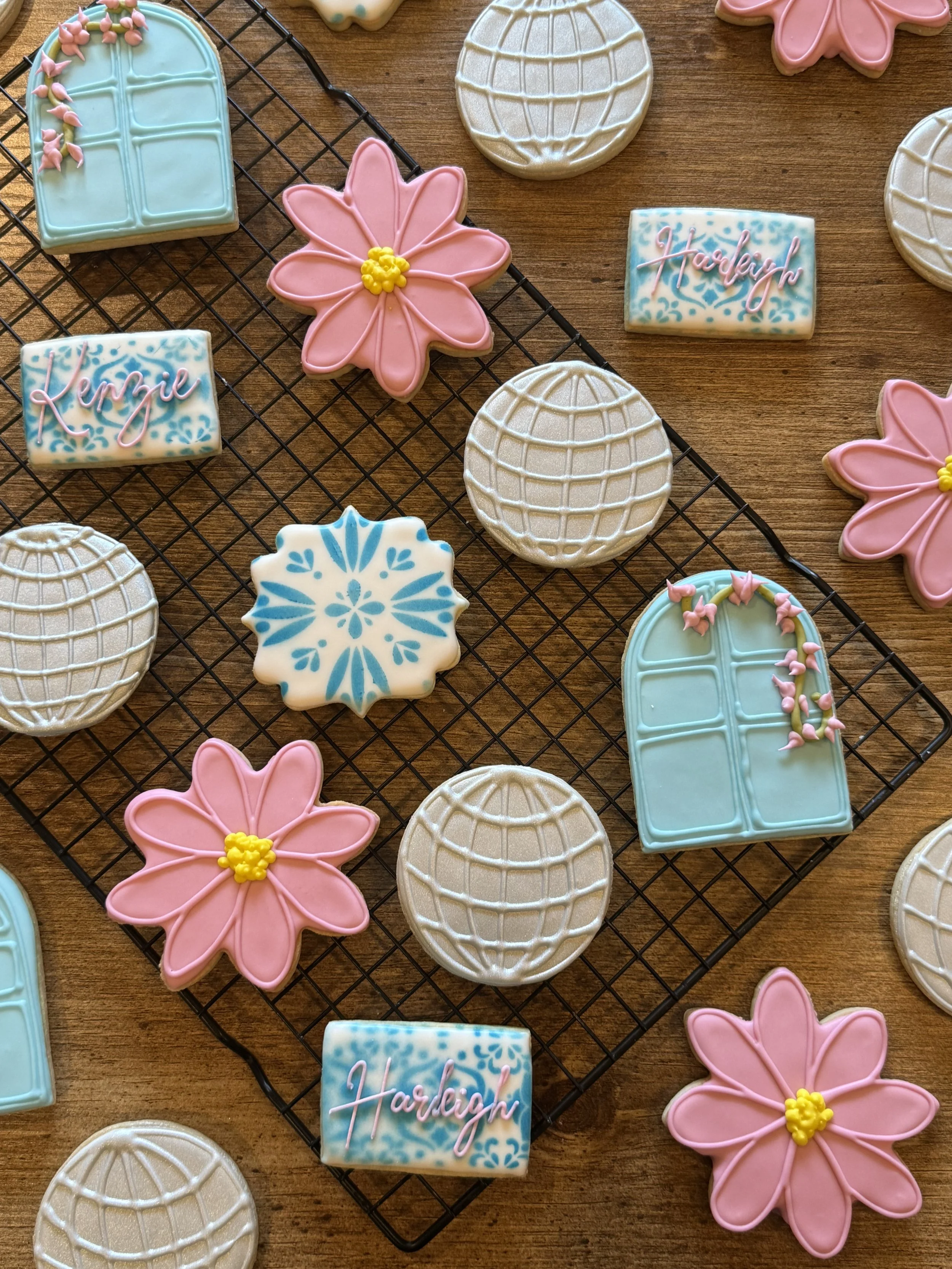 Decorative cookies on a cooling rack and wooden surface, including pink flowers, blue snowflakes, globe designs, and cookies inscribed with the name 'Haleigh' in pink icing.