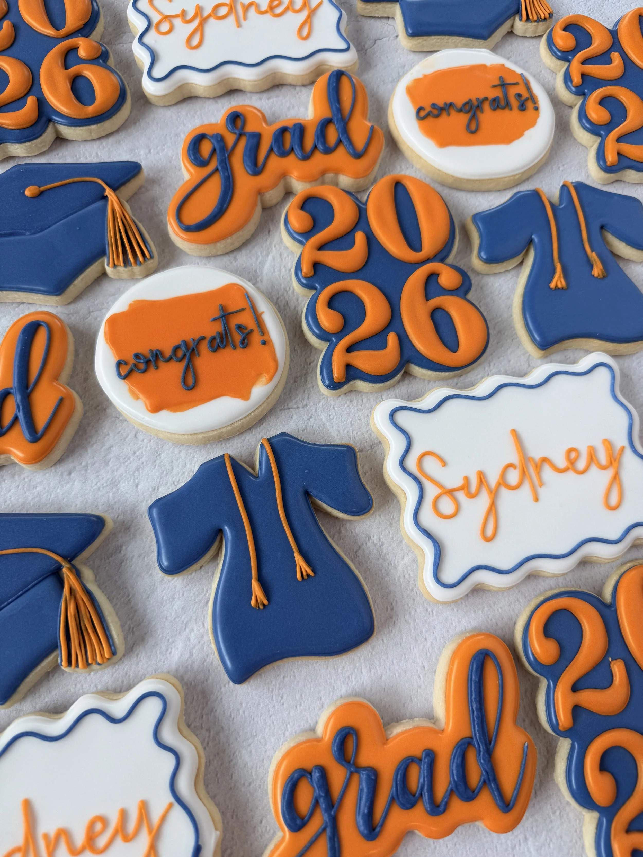 Collection of decorated cookies with graduation-themed messages and designs in blue, orange, and white, including graduation caps, the word 'grad', 'congrat', 'thank you', and the year 2026.