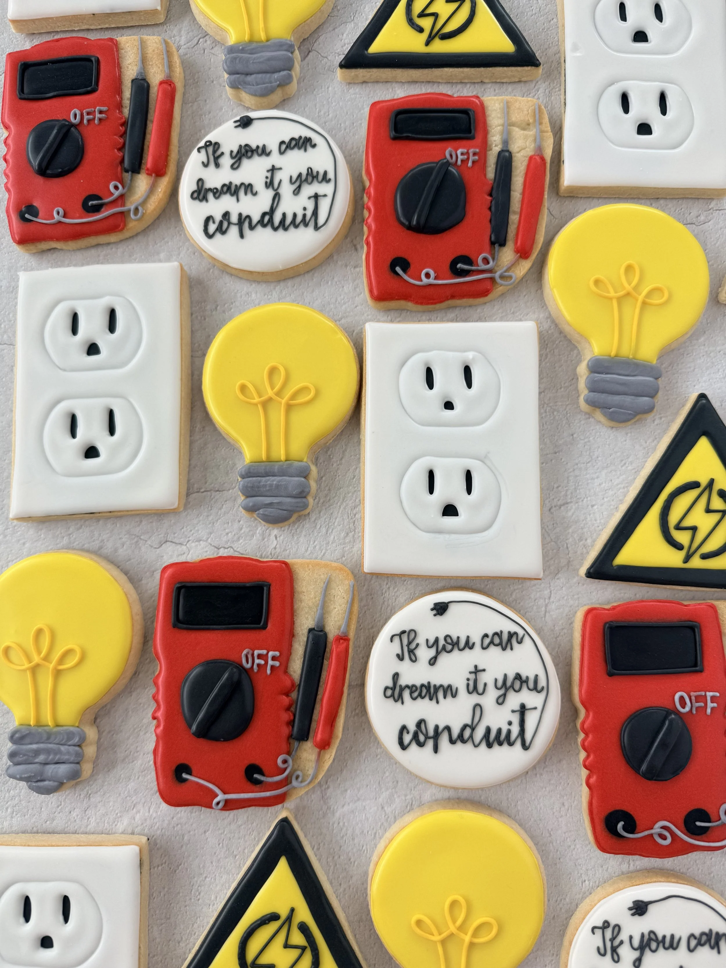 Cookies decorated with electrical safety themes, including light bulbs, power outlets, electrical hazard signs, and electrical switches. Some cookies also feature writing, such as a phrase about dreaming it you can conduct it.