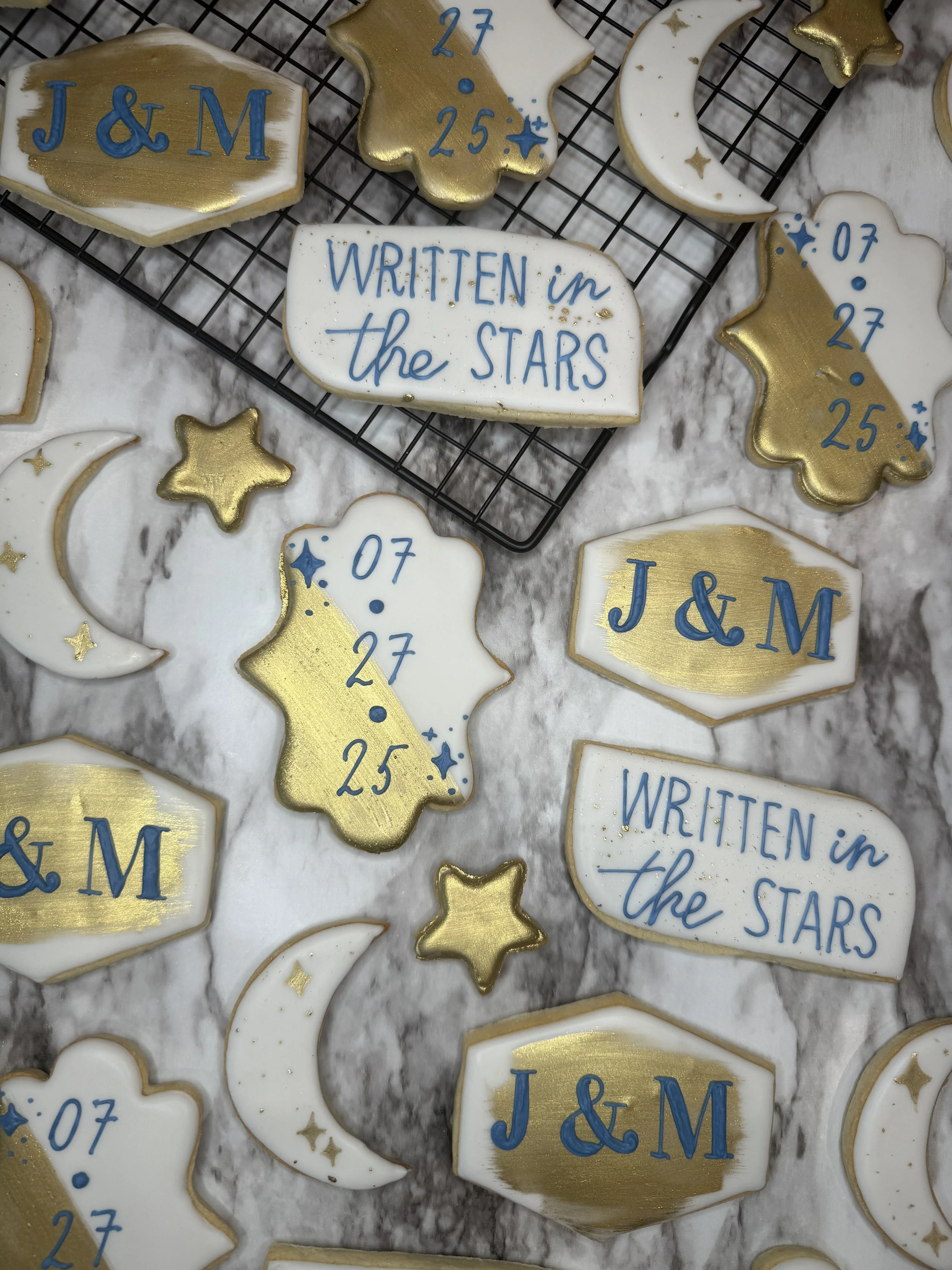 Decorative cookies with celestial themes, including a crescent moon, stars, and clouds, with gold and blue accents, some with inscriptions like "J & M," "07 27 25," and "Written in the stars."
