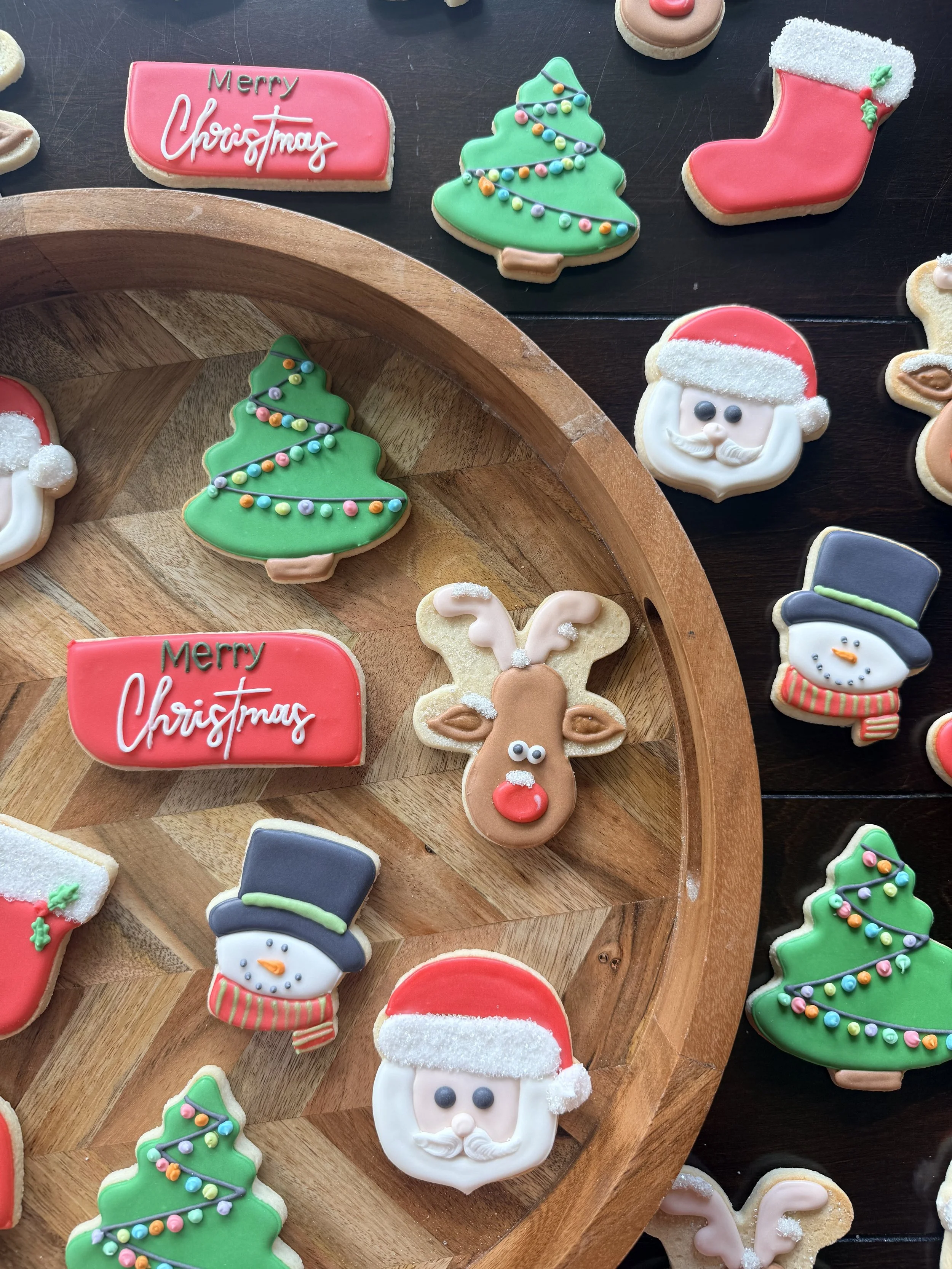 Christmas-themed cookies shaped like Christmas trees, Santa Claus, reindeer, snowmen, a hat, a mitten, a nose, and a 'Merry Christmas' sign, decorated with colorful icing and sprinkles, placed on a wooden and dark table.