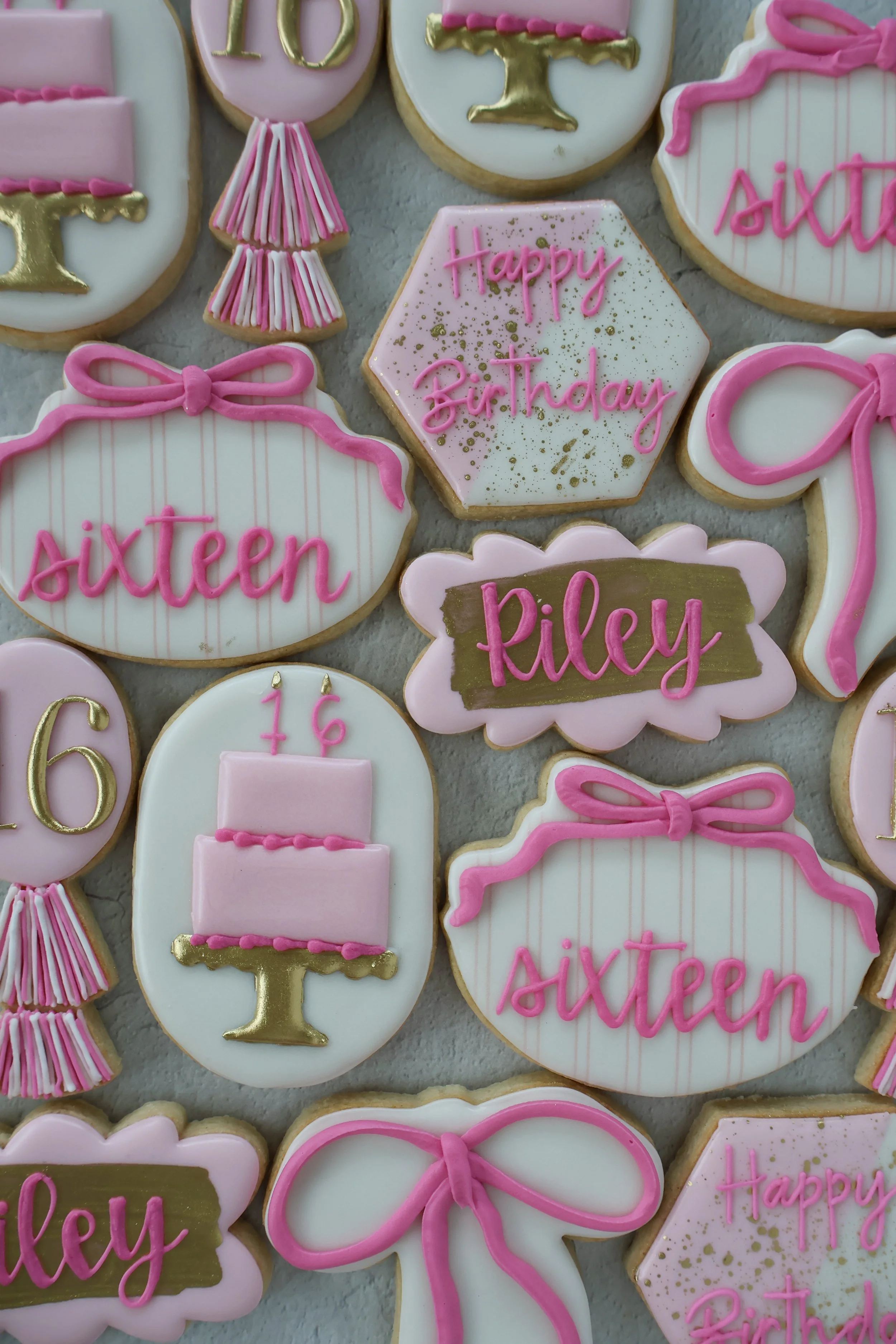 Collection of decorated cookies for a birthday celebration. The cookies are decorated in pink, white, and gold, with messages like "Happy Birthday" and names such as "Riley." Some cookies feature bows, a tiered cake, and the number 16, indicating a 1