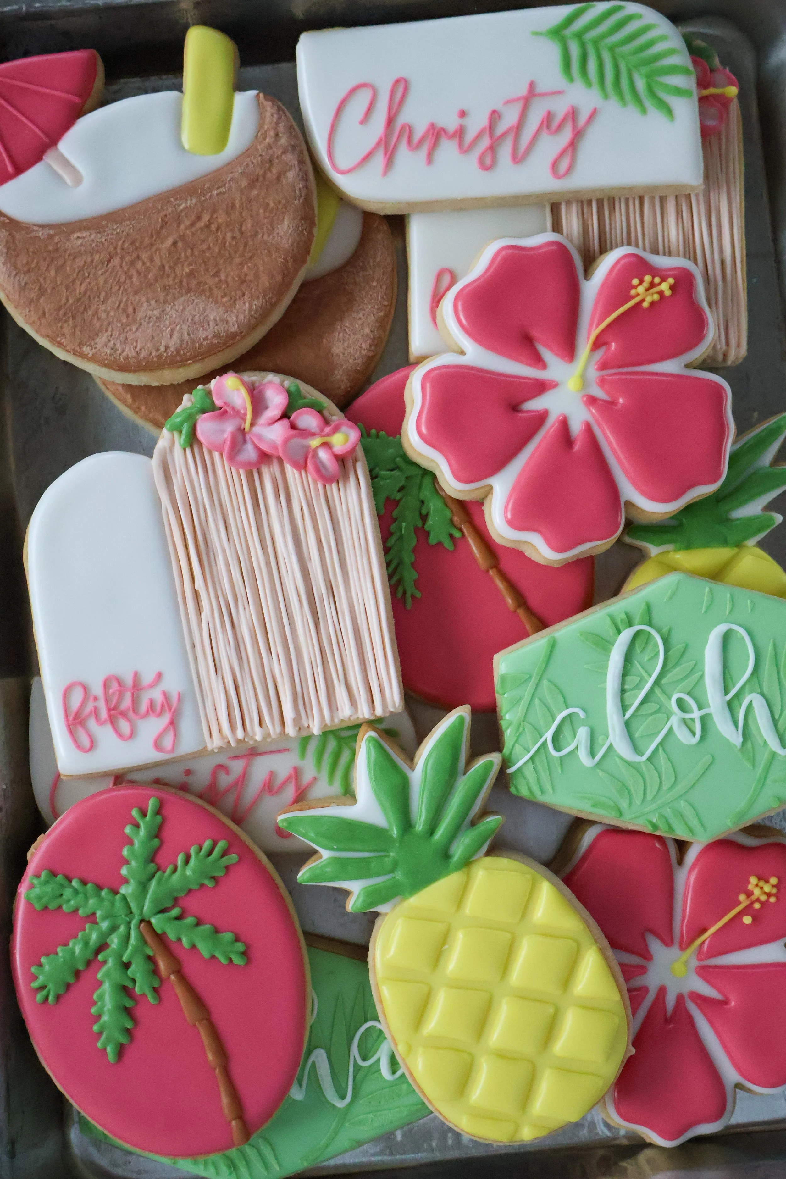 Decorated sugar cookies with tropical and birthday themes, including pineapple, hibiscus flower, palm tree, and personalized cookie with the name 'Christy'.