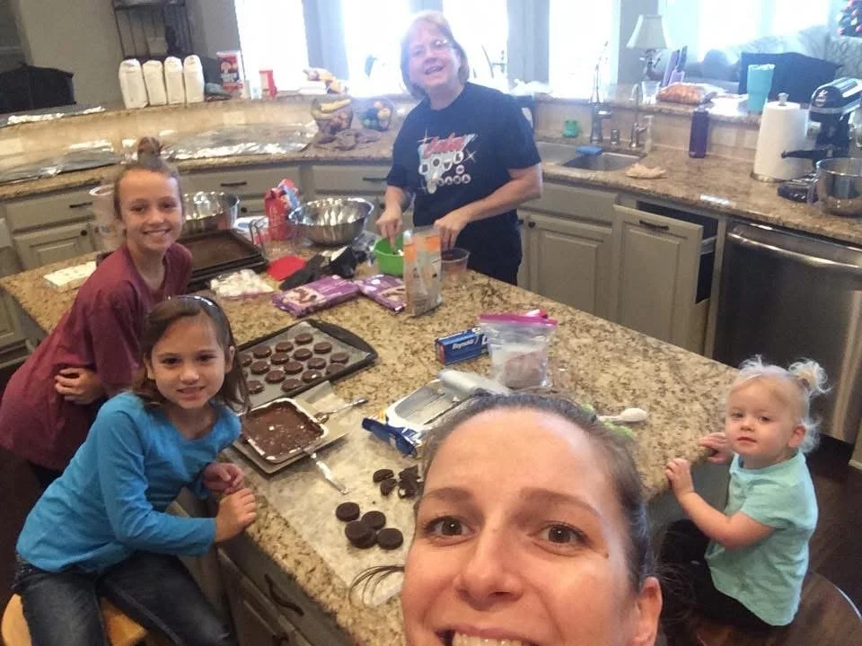 Family in a kitchen baking cookies together, with cookie dough, chocolate cookies, and baking supplies on the counter.