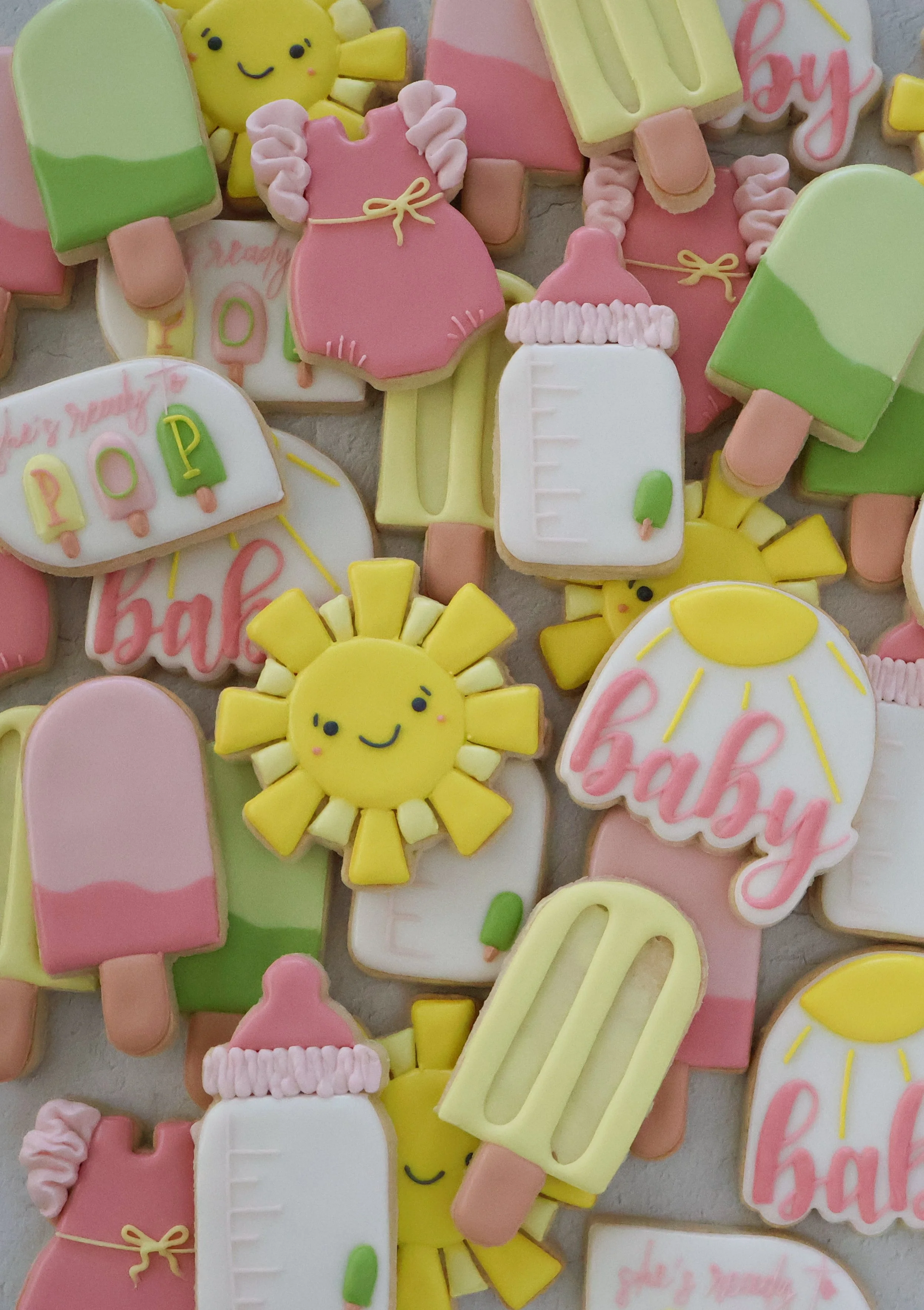 Collection of colorful baby-themed cookies shaped like popsicles, suns, milk bottles, and onesies. Some cookies have decorative writing that says 'Baby' and 'Happy' in pink and green icing.