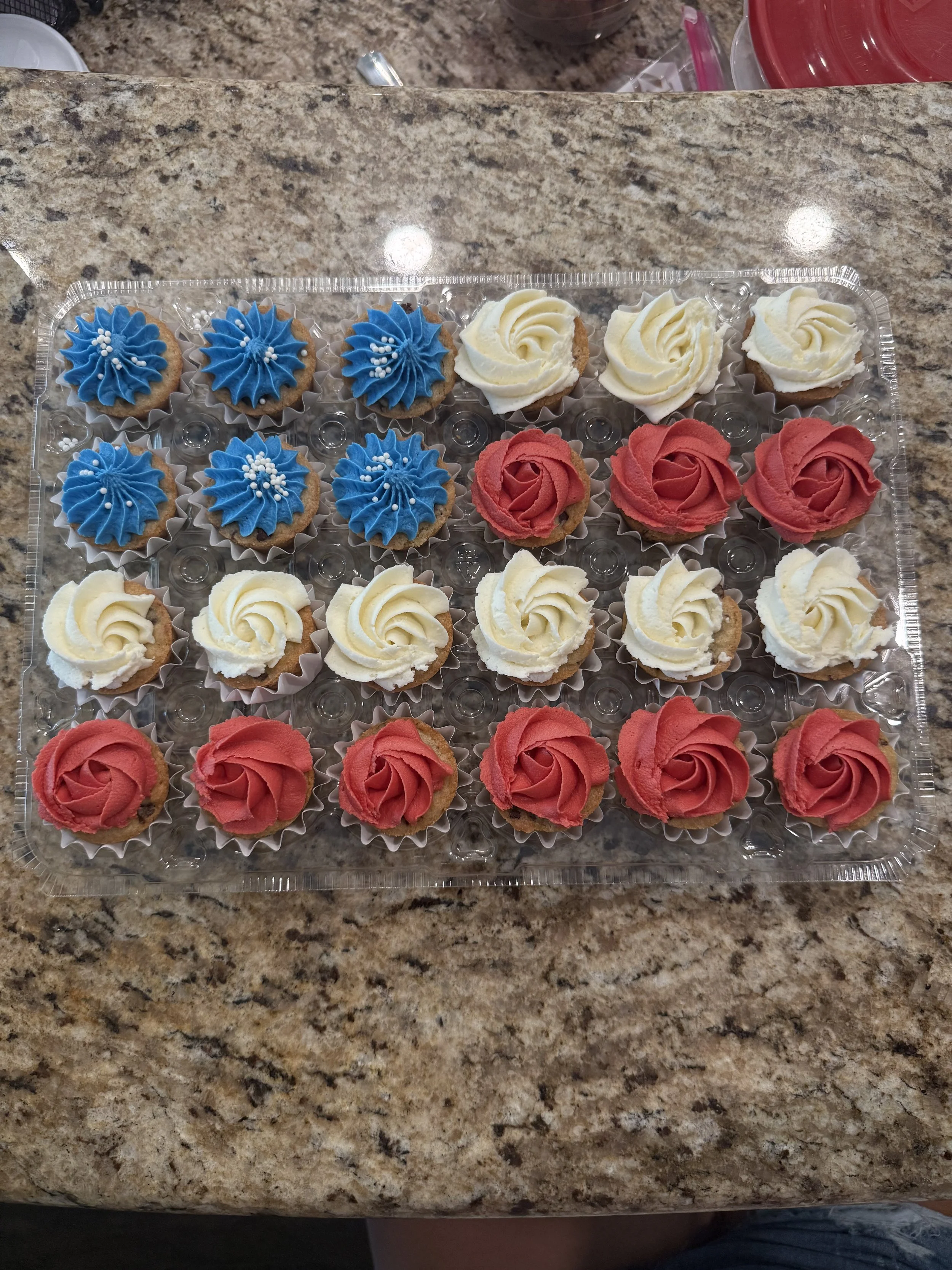 A plastic tray of decorated cupcakes on a marble countertop, with blue, white, and red frosting, topped with small white sprinkles.