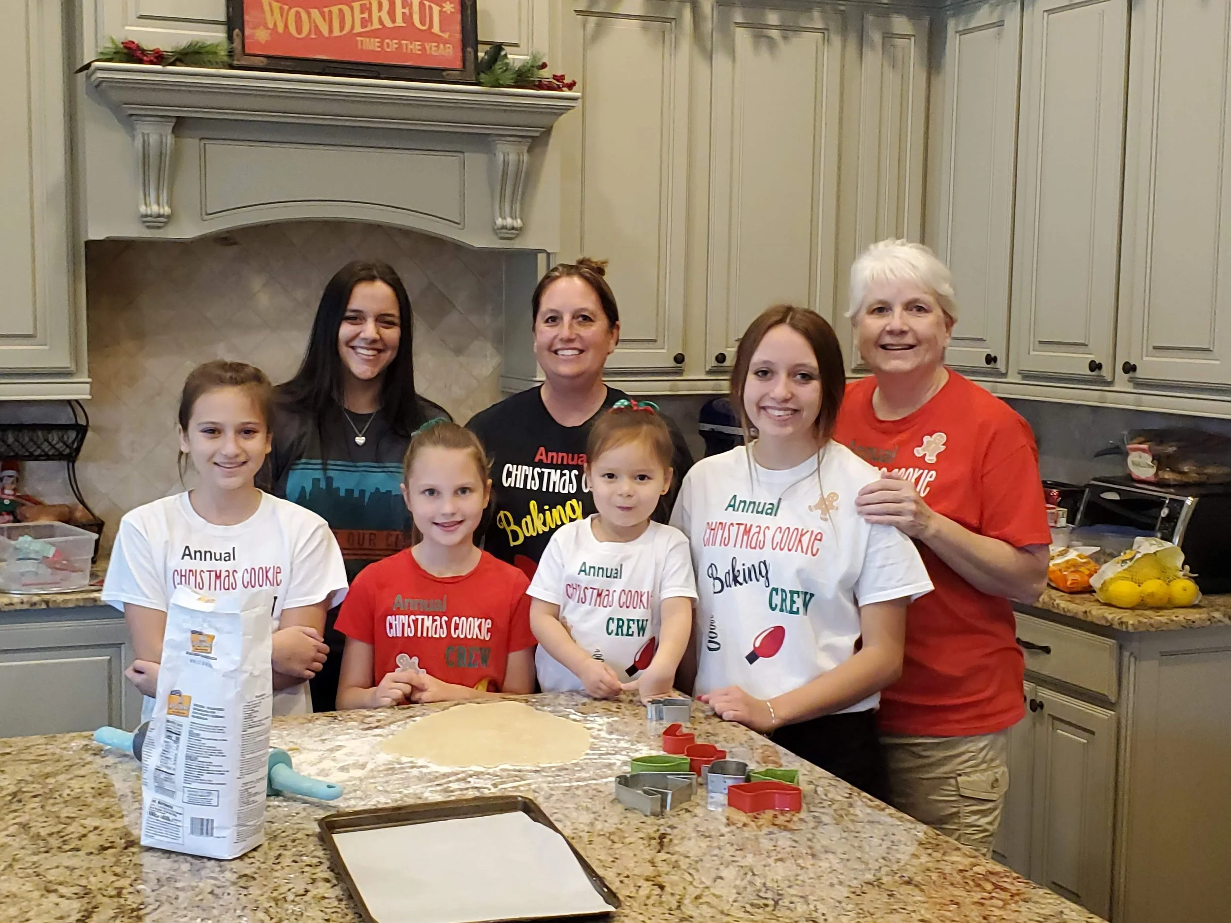 Group of people, including children and adults, gathered in a kitchen participating in a holiday cookie decorating activity. The children are wearing shirts that read 'Annual Christmas Cookie Baking Crew,' and the adults are smiling, with one wearing