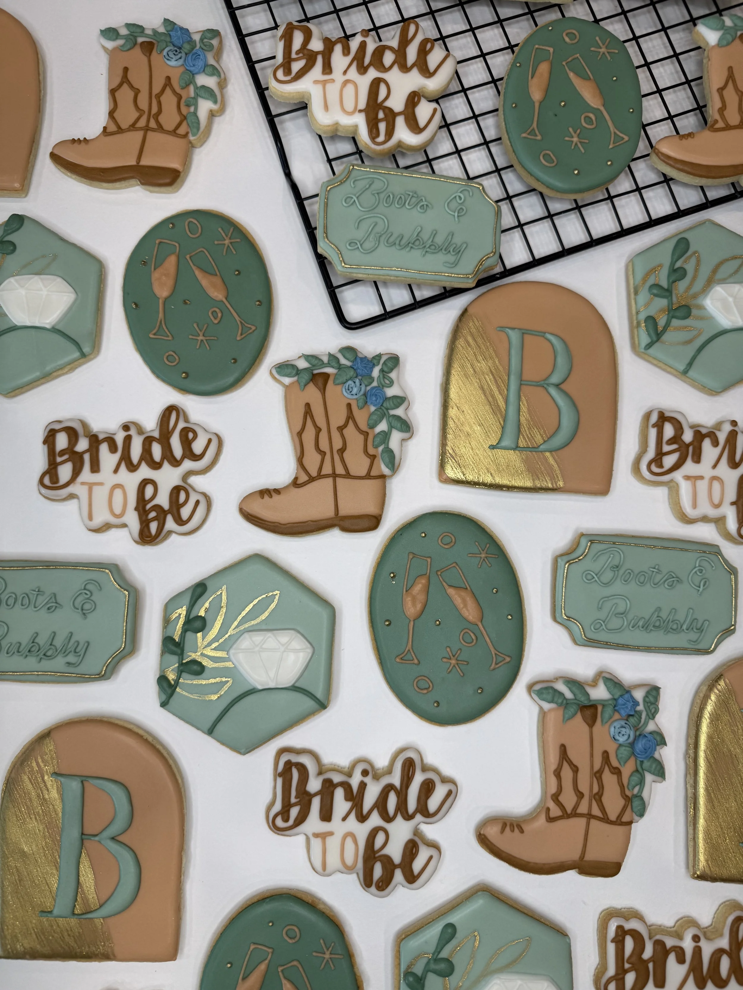 Decorative cookies with wedding and celebration themes, including cowboy boots with blue flowers, champagne glasses, and text reading 'Bride to Be' and 'Boots & Bubbly' in pastel colors with gold accents, arranged on a white surface and some on a bla
