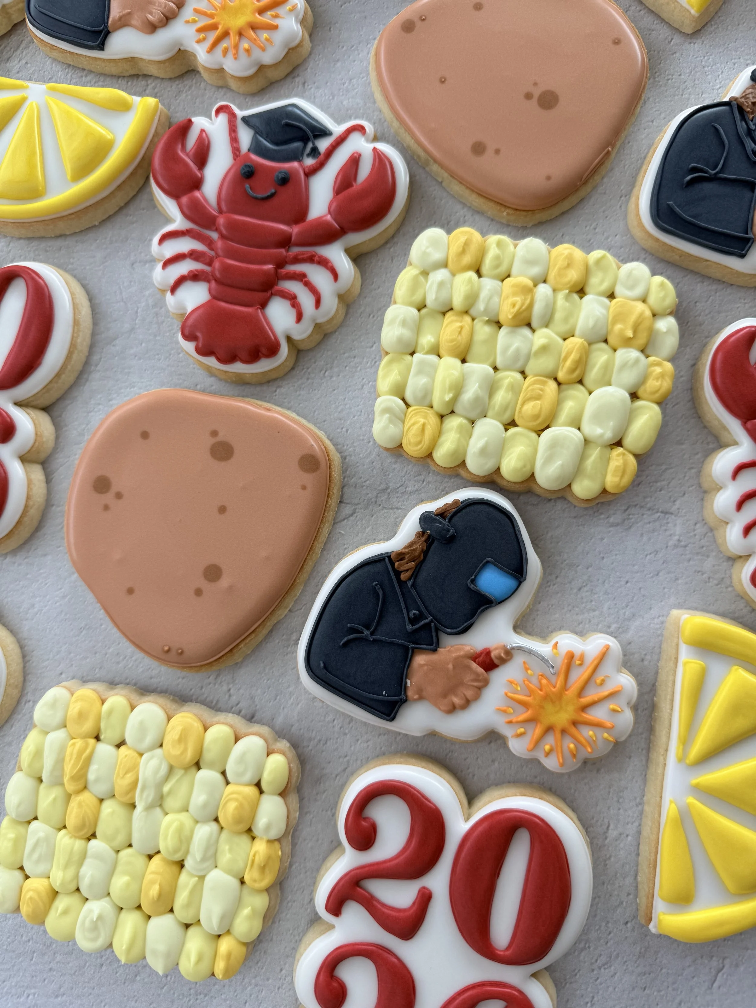 Decorated sugar cookies with various designs, including a red lobster with a graduation cap, yellow and white corn, a brown hot dog bun, black shoes, a love heart, and a red number 20.