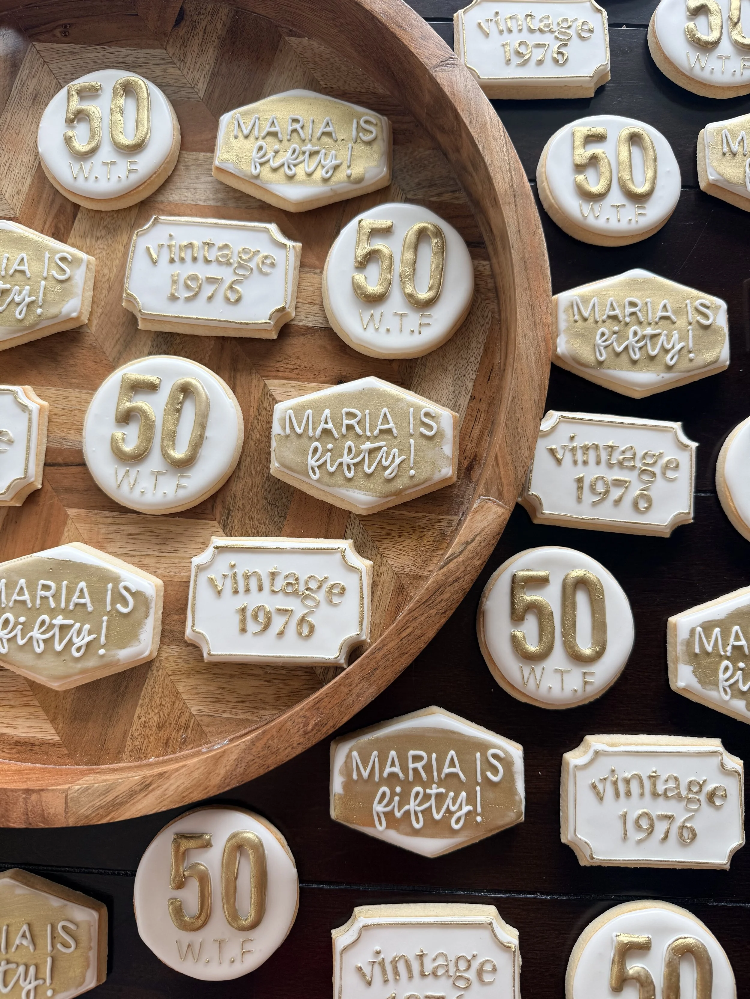 Cookies decorated with gold and white icing, celebrating Maria's 50th birthday, with some cookies reading "50 W.T.F," "Maria is fifty!," and "vintage 1976".