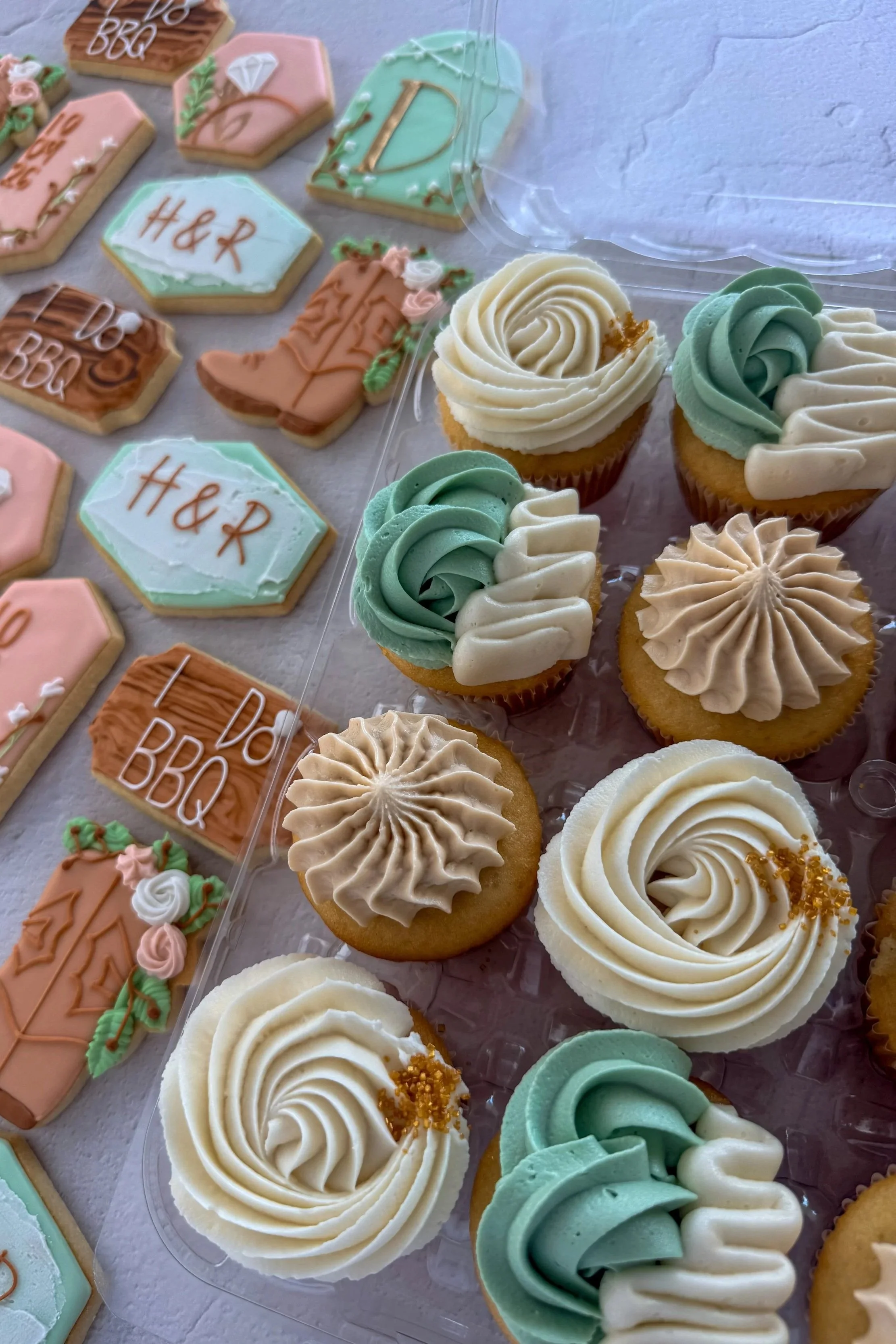 Assorted decorated cookies and cupcakes, including cookies with 'H&R' and 'Dare BBQ' written on them, and cupcakes with swirl frosting in white, green, and beige, some with gold sprinkles.