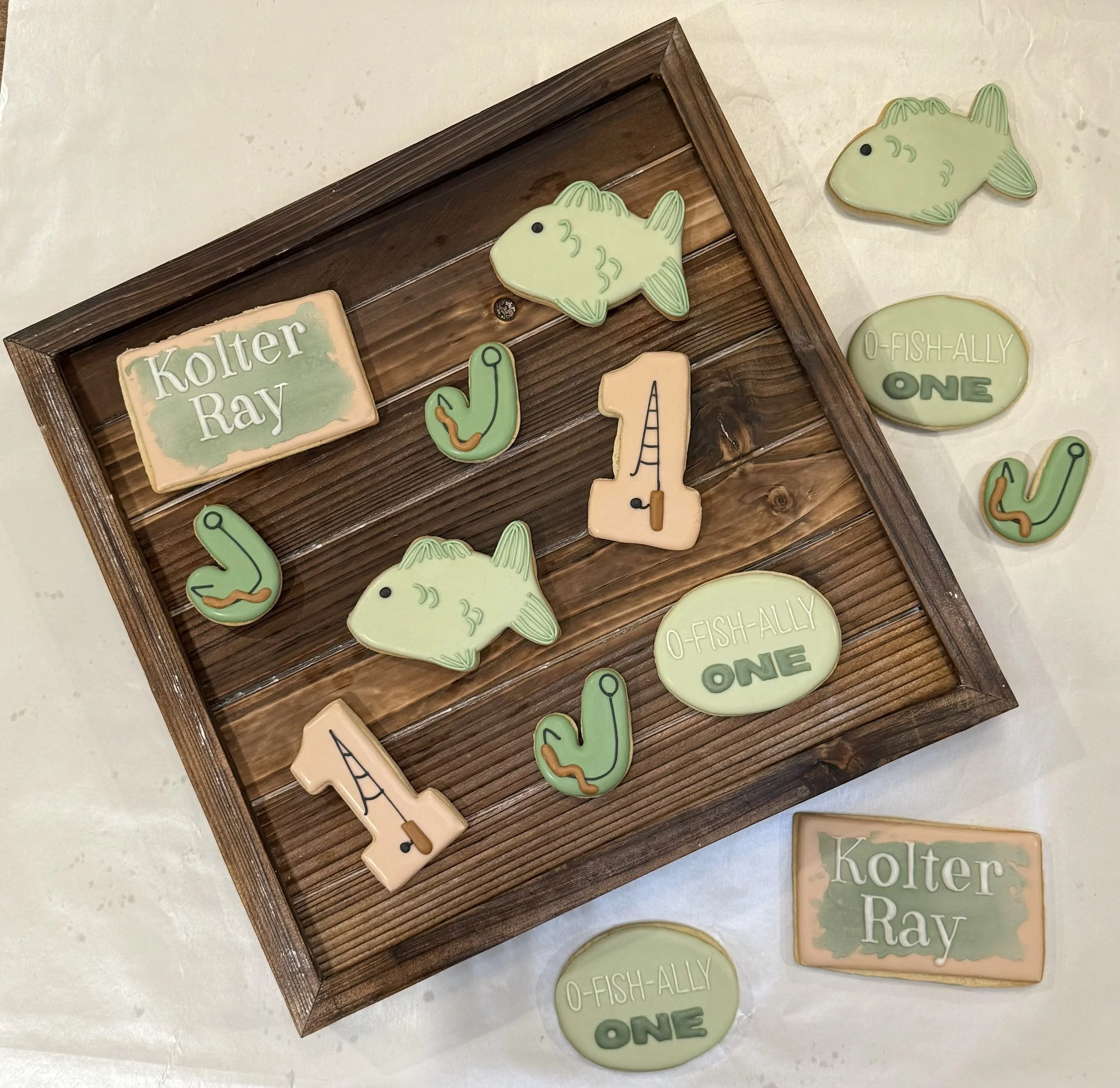 Decorative cookies shaped like a fish, a cactus, and other shapes, with text reading 'Kolter Ray' and 'O-FISH-ALLY ONE', displayed on a wooden tray and white surface.