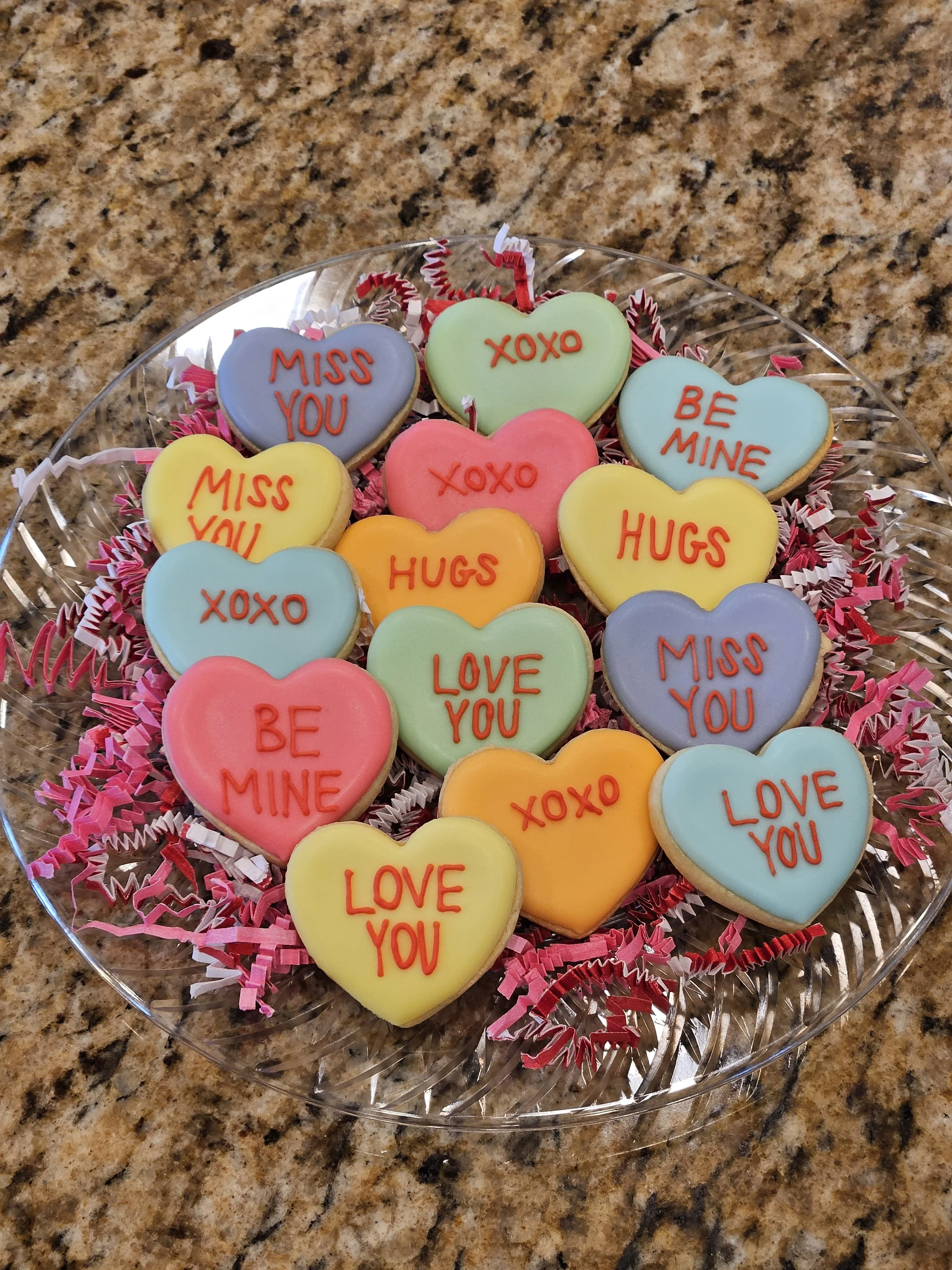 Decorated heart-shaped cookies with messages like "Miss You," "XOXO," "Be Mine," and "Hugs" on a clear glass tray, with pink and white shredded paper for decoration, on a granite countertop.