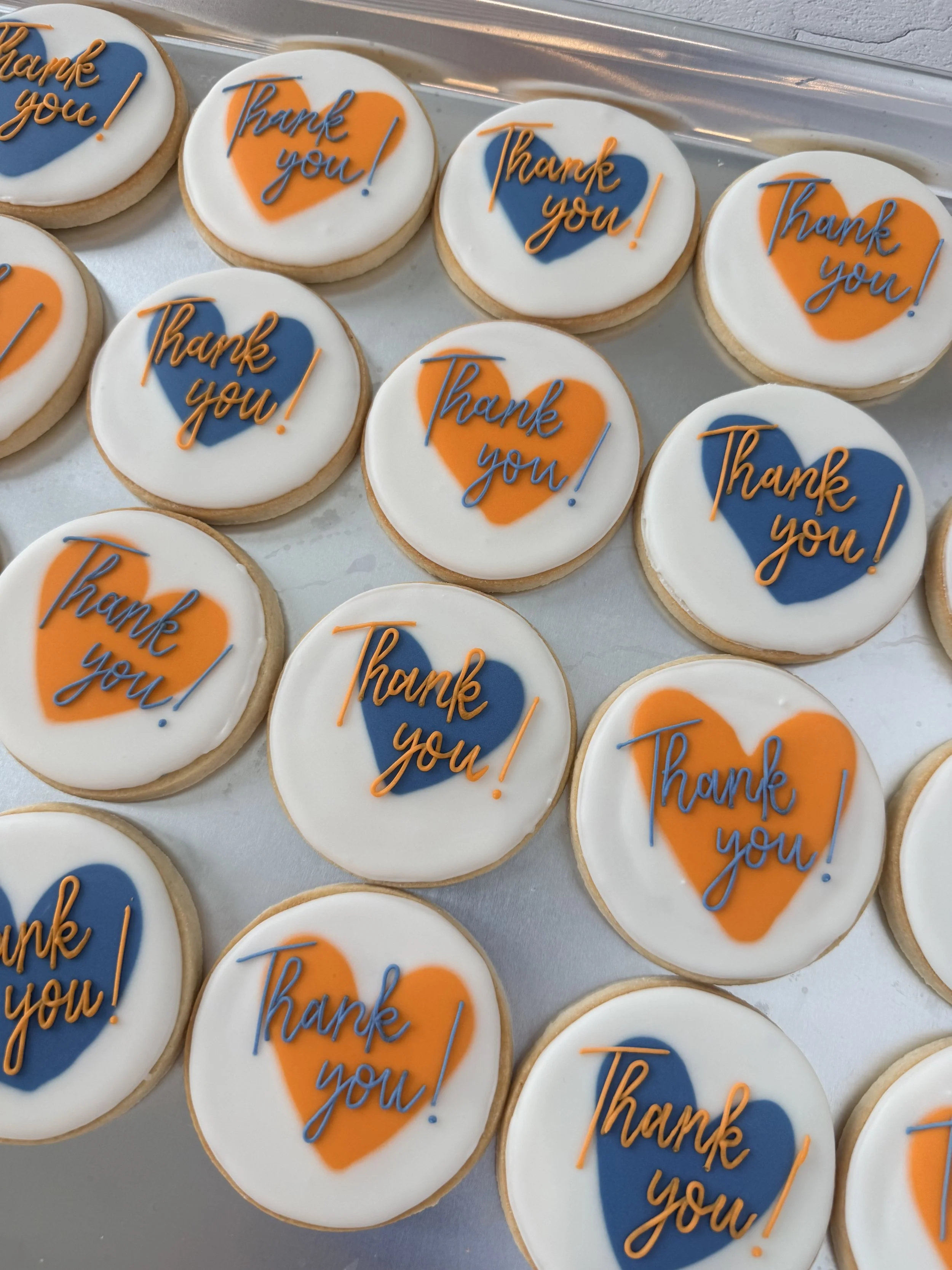 Cookies decorated with edible icing, each featuring a heart in orange and blue with the words 'Thank you!' written in orange cursive icing.