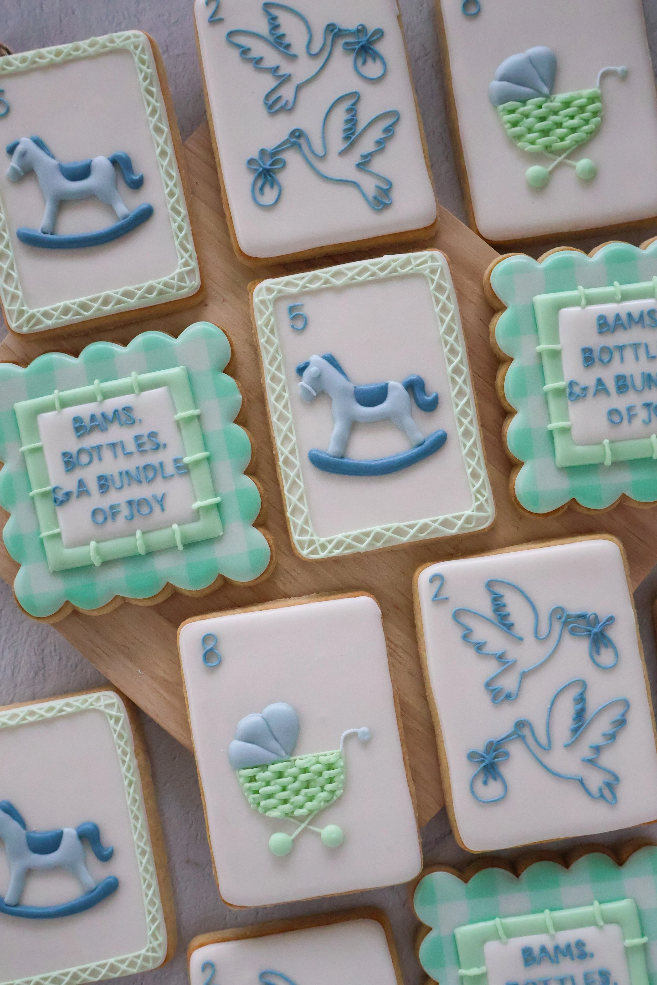 Decorative cookies with baby-themed designs including rocking horses, storks, baby strollers, and a basket of baby bottles and toys in pastel colors, arranged on a wooden surface.