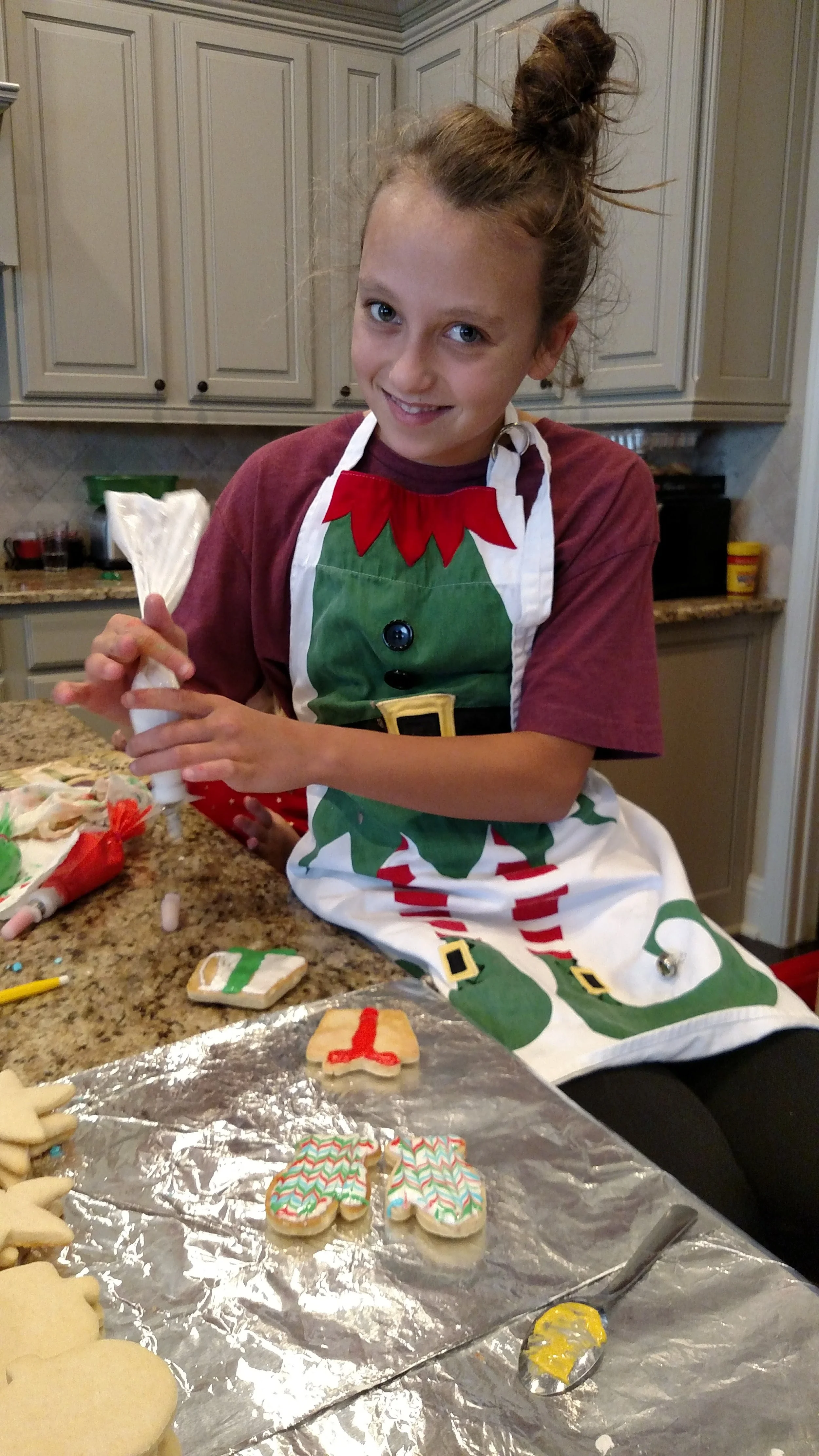 A girl wearing a Christmas-themed apron is decorating holiday cookies in a kitchen. She has light brown hair tied in a bun and is smiling while holding a piping bag.