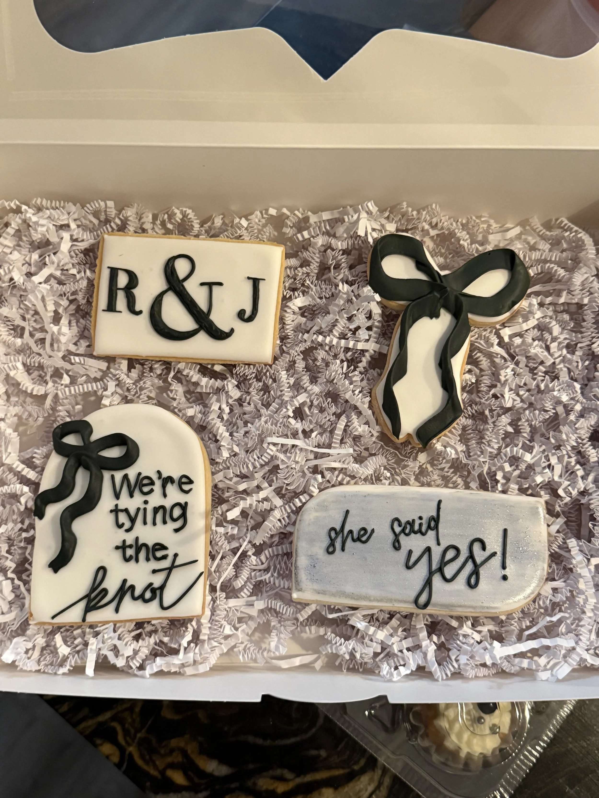Decorative cookies with black and white icing, including one with the initials 'R & J' and another with the phrase 'We're tying the knot' along with a cookie shaped like a bow tie, all placed on shredded paper in a box.