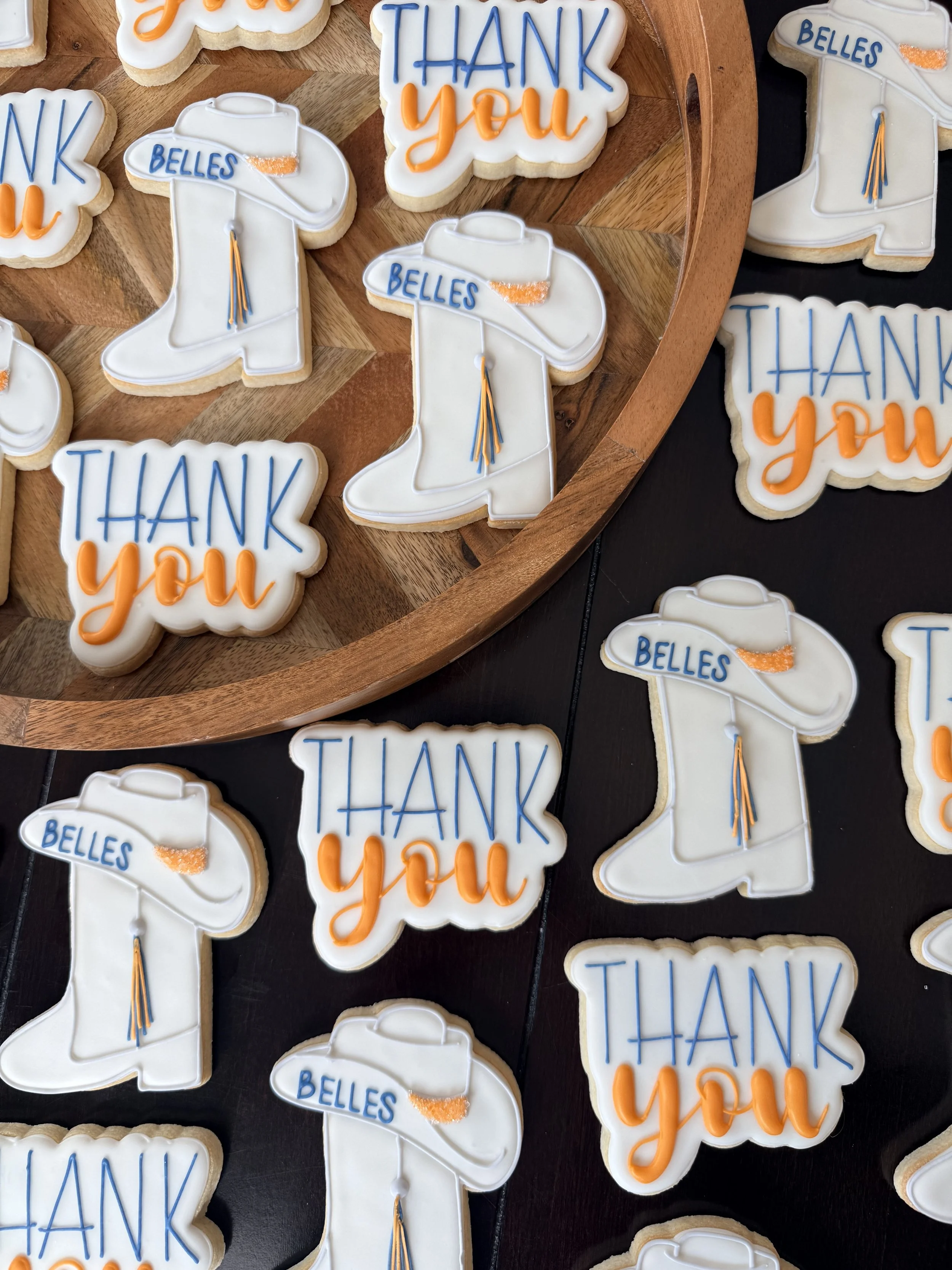 Decorative cookies with white icing shaped like hats, featuring blue and orange writing that says "Belles" and "Thank you," with some cookies shaped like graduation caps with tassels.