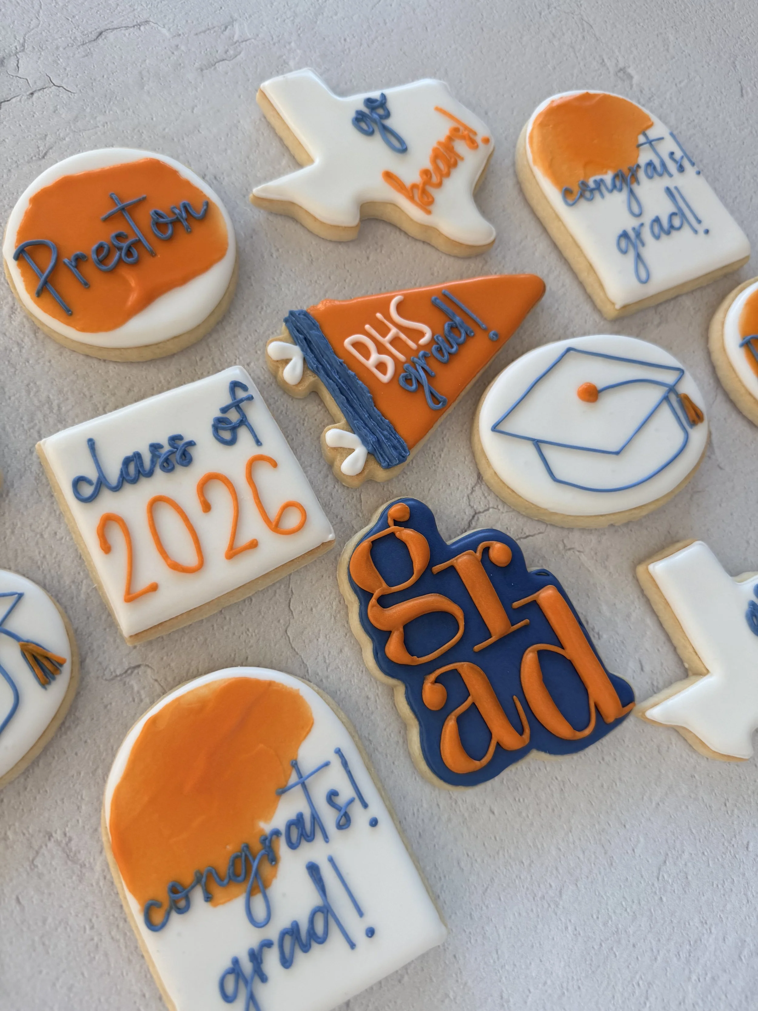 Collection of decorated cookies celebrating high school graduation with various phrases and graduation symbols in orange, white, and blue icing.