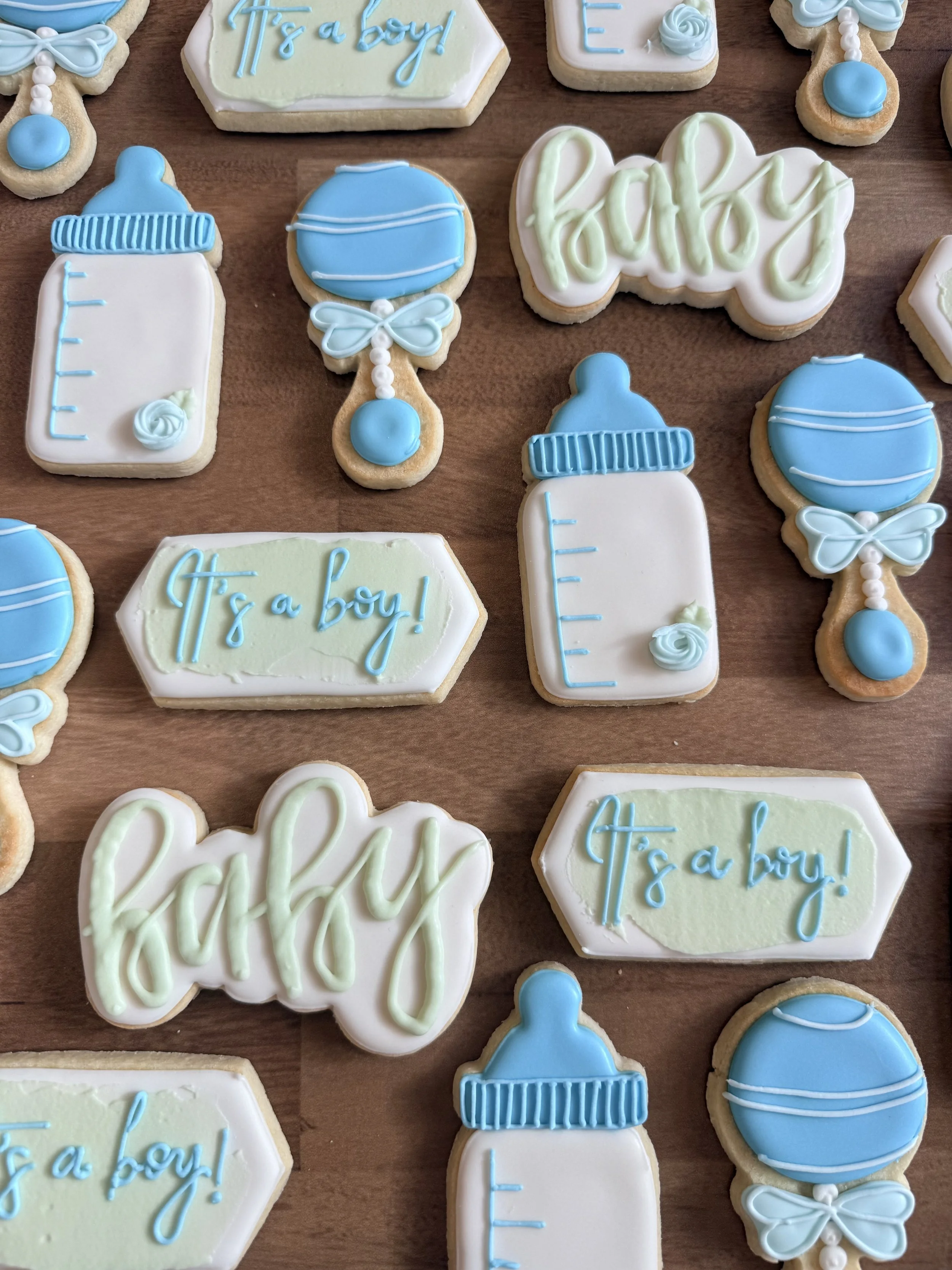 Decorated cookies with baby-themed designs and messages, featuring blue and white icing. Some cookies have baby bottles, pacifiers, and the phrase 'It's a boy!' written on them.