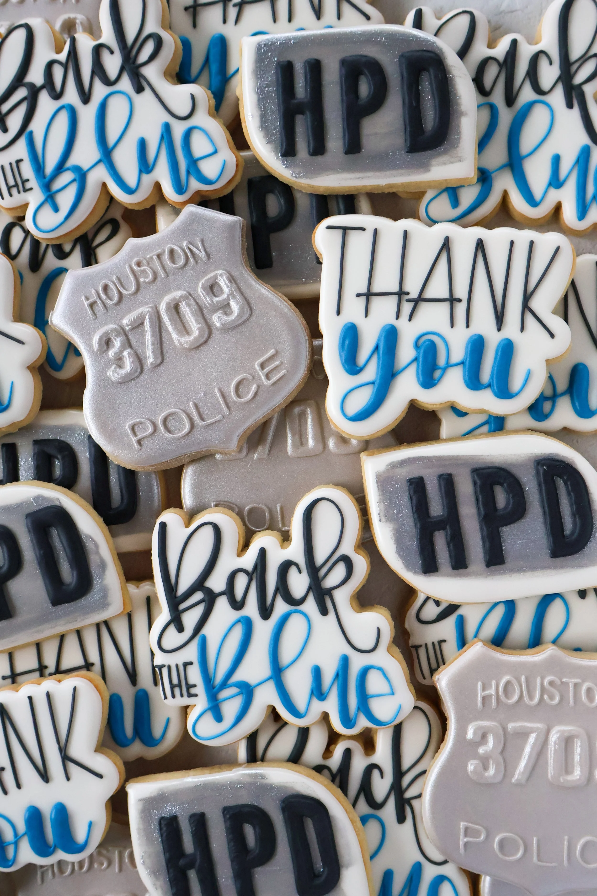 Decorative cookies with messages like 'Thank You' and 'Happy' in various colors and designs, including a silver police badge-shaped cookie with 'Houston Police 3109' embossed on it.