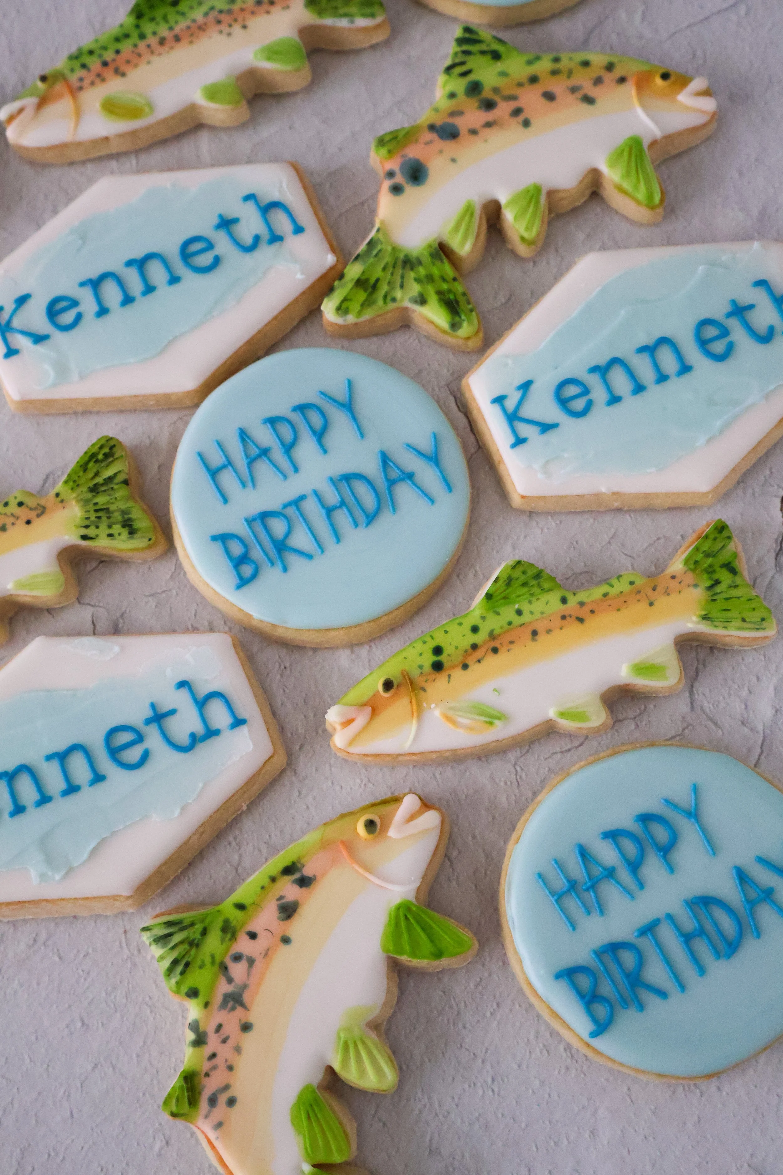Decorated cookies shaped like fish, with some cookies saying 'Happy Birthday' and others with the name 'Kenneth' written on them, displayed on a flat surface.