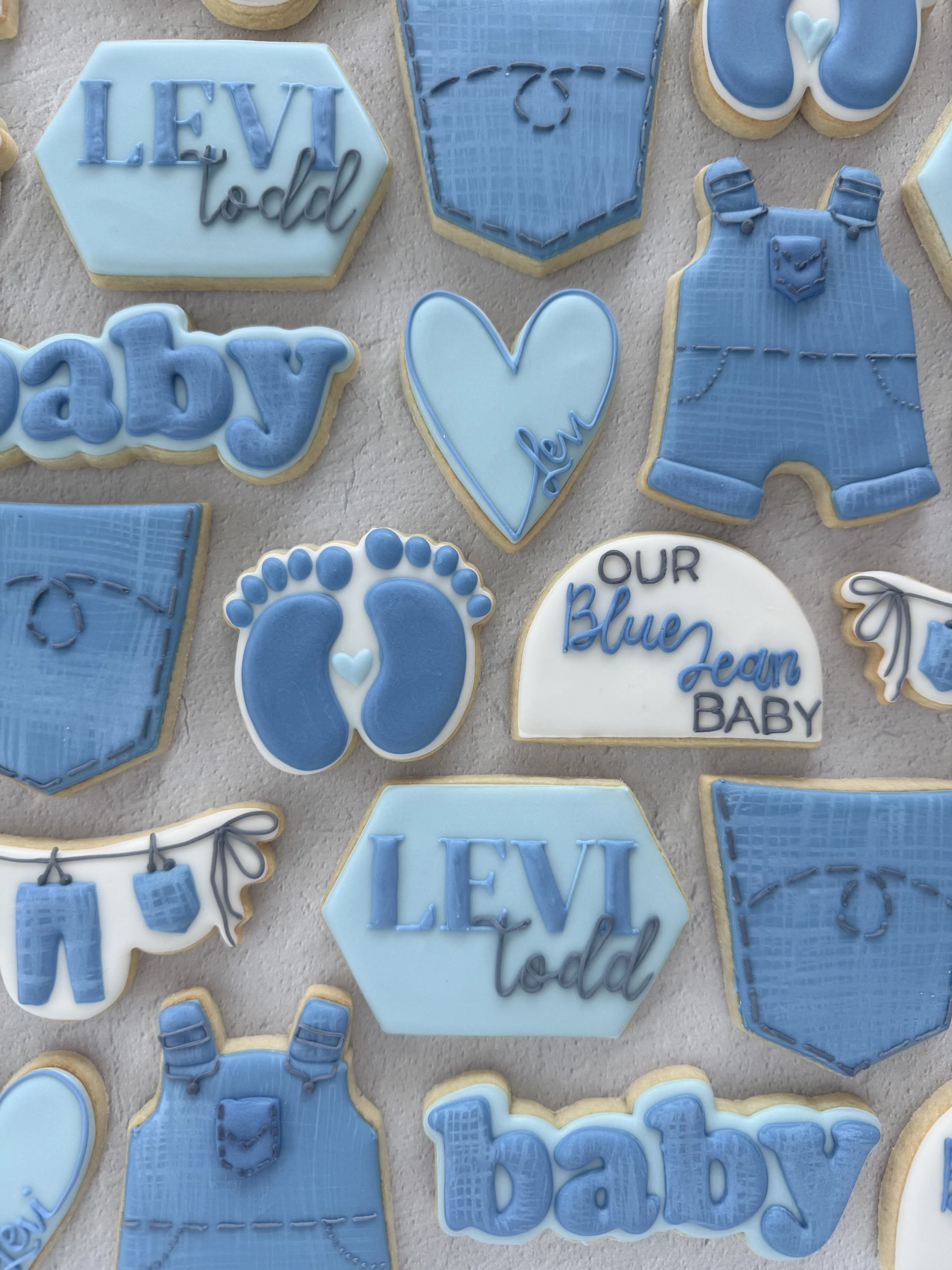 Set of baby shower cookies decorated with blue, white, and gray icing, featuring words like 'baby,' 'Levi,' 'dad,' and images of footprints, bibs, and clothing.