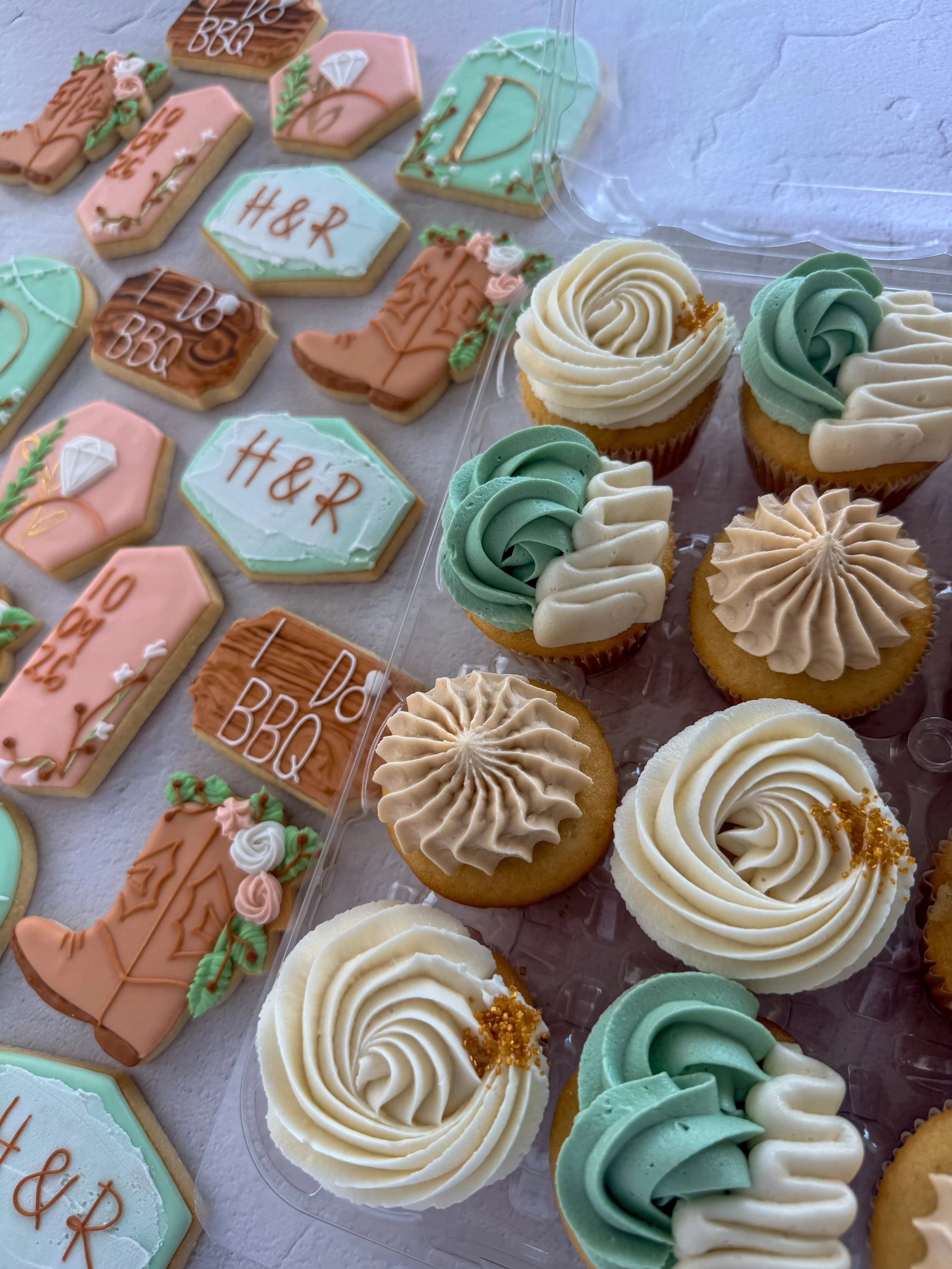 Assorted decorated cookies and cupcakes with pastel icing and themed decorations, including monogrammed cookies and a cowboy boot cookie, arranged on a white surface.
