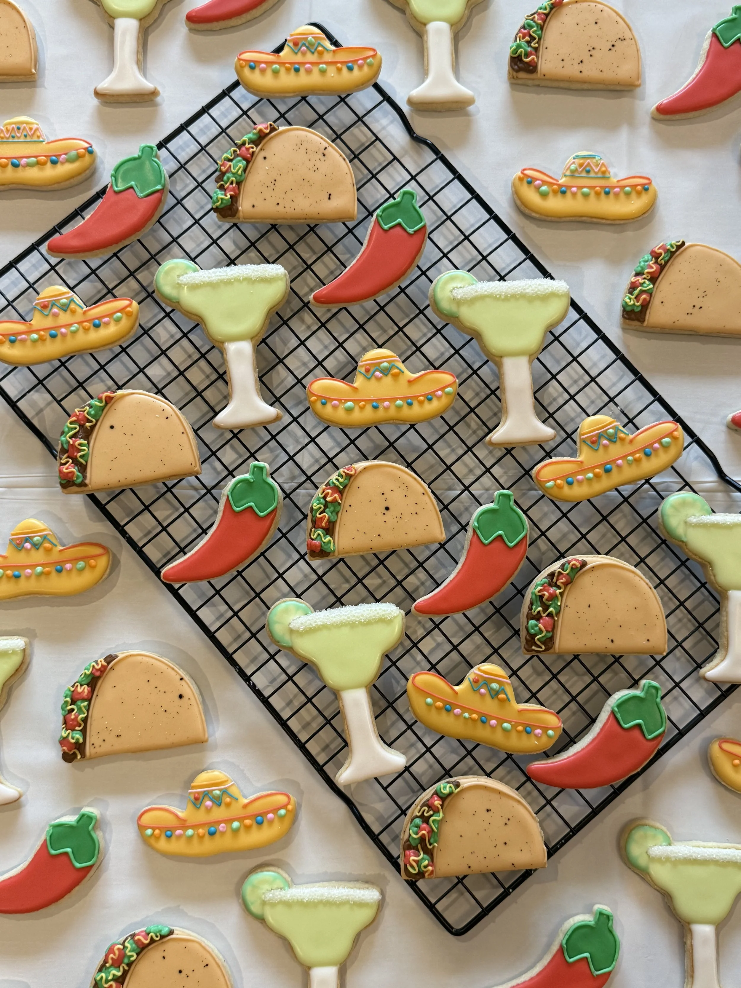 Decorated cookies in the shape of Mexican dishes, including tacos, chili peppers, margarita glasses, and sombreros, arranged on a wire rack and parchment paper.