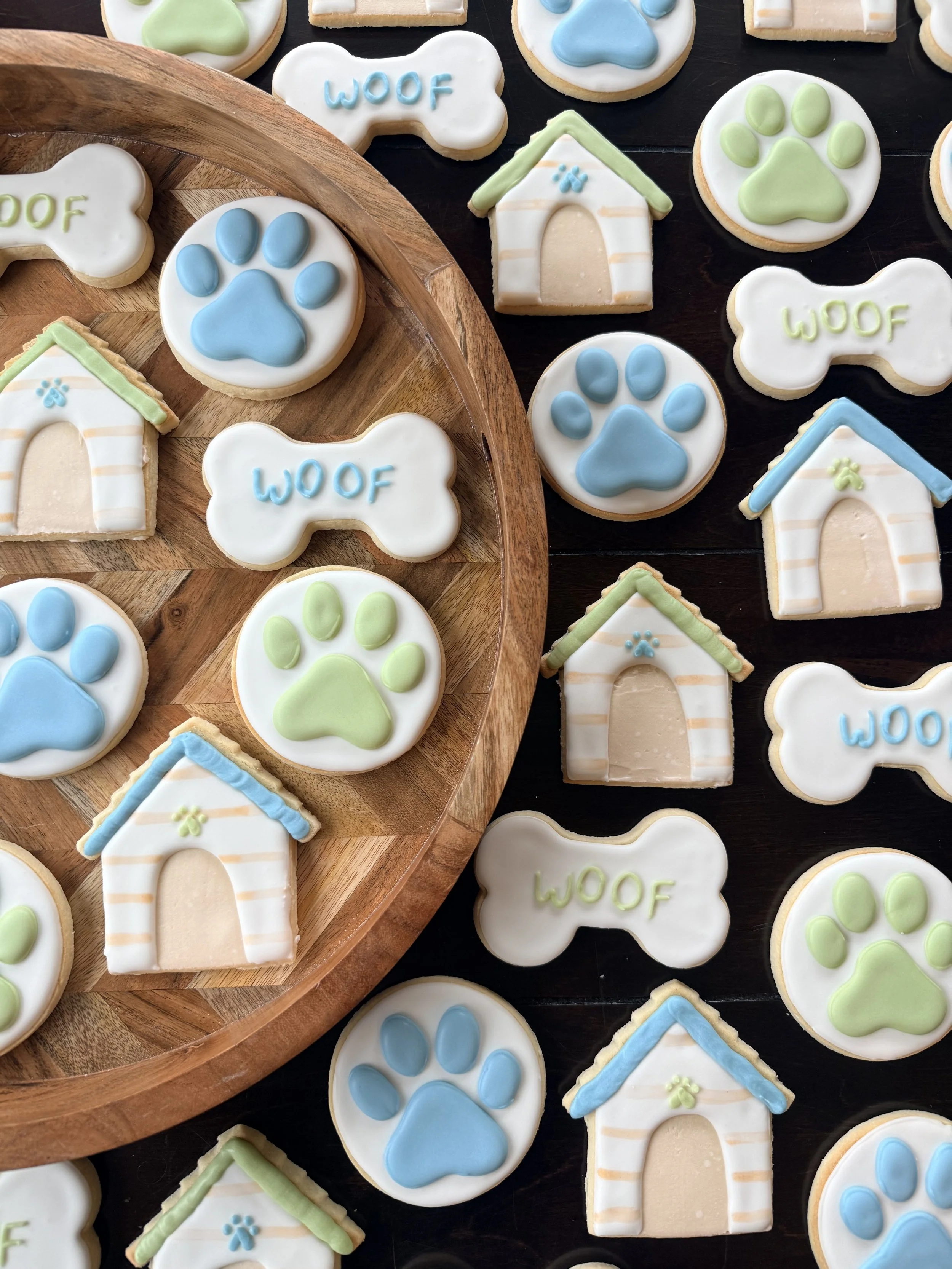 Decorated dog-shaped cookies, paw print cookies, and house-shaped cookies with pastel icing in blue, green, and white colors displayed on a black surface and a wooden tray.