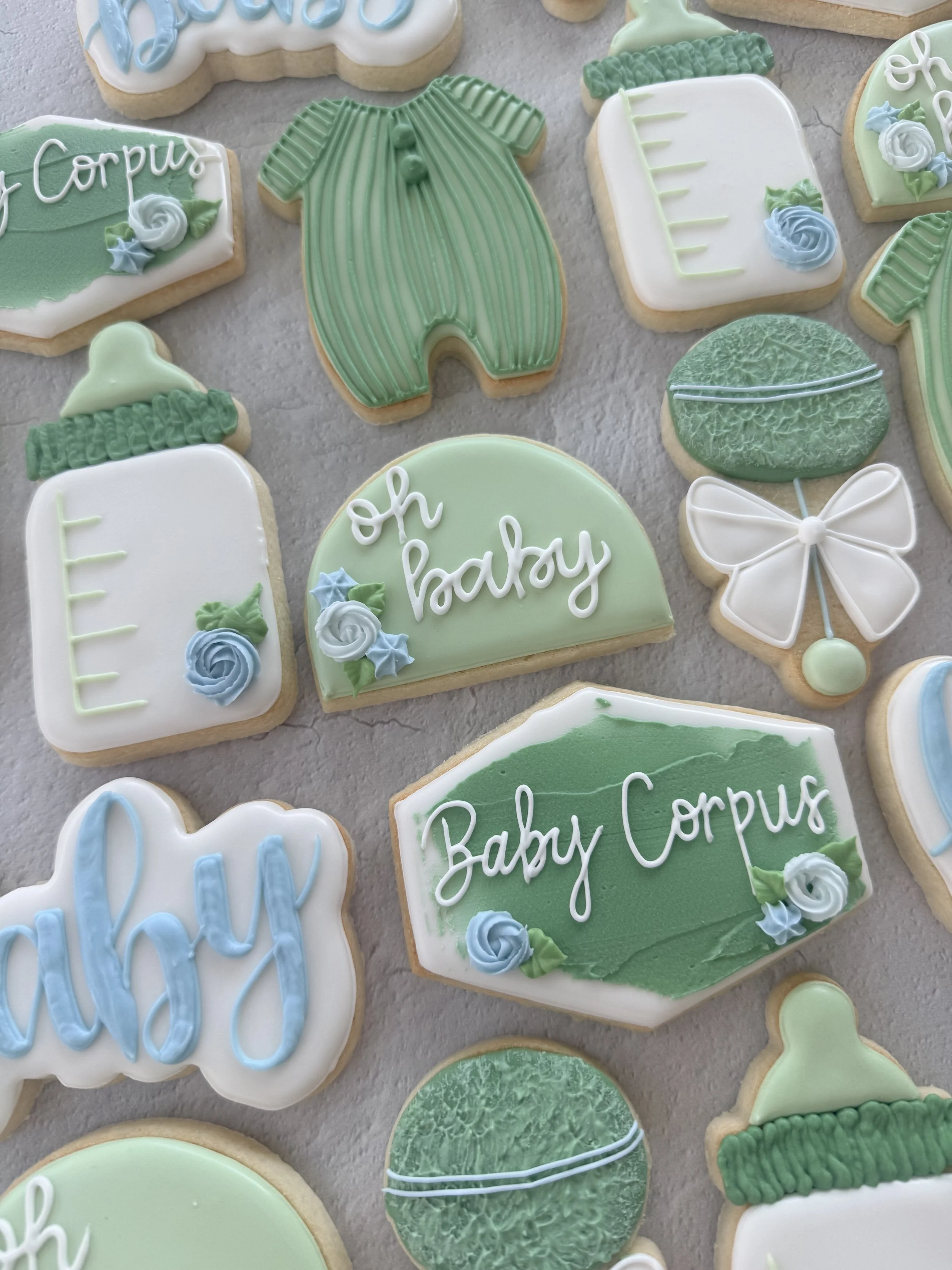 A collection of cookies decorated with pastel-colored icing, some with baby shower themes, including ones with baby clothes, baby bottles, and bouquets of flowers, in shades of green, blue, and white.