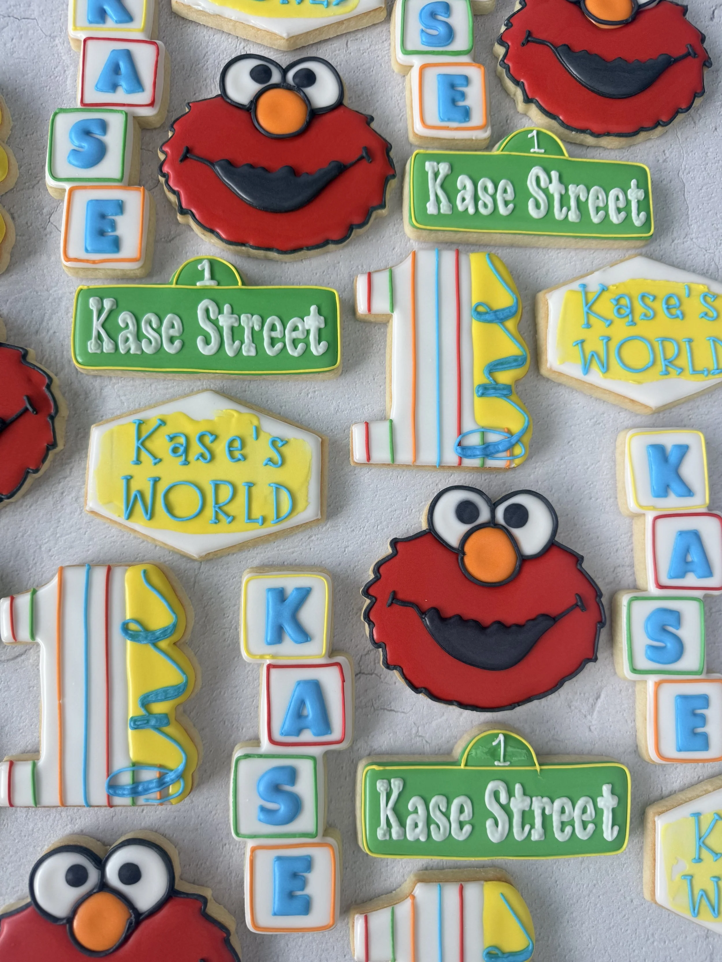 Colorful decorated cookies with Sesame Street theme, featuring Elmo's face, street signs with 'Kase Street' and 'Kase's World,' and a number 1 party hat.