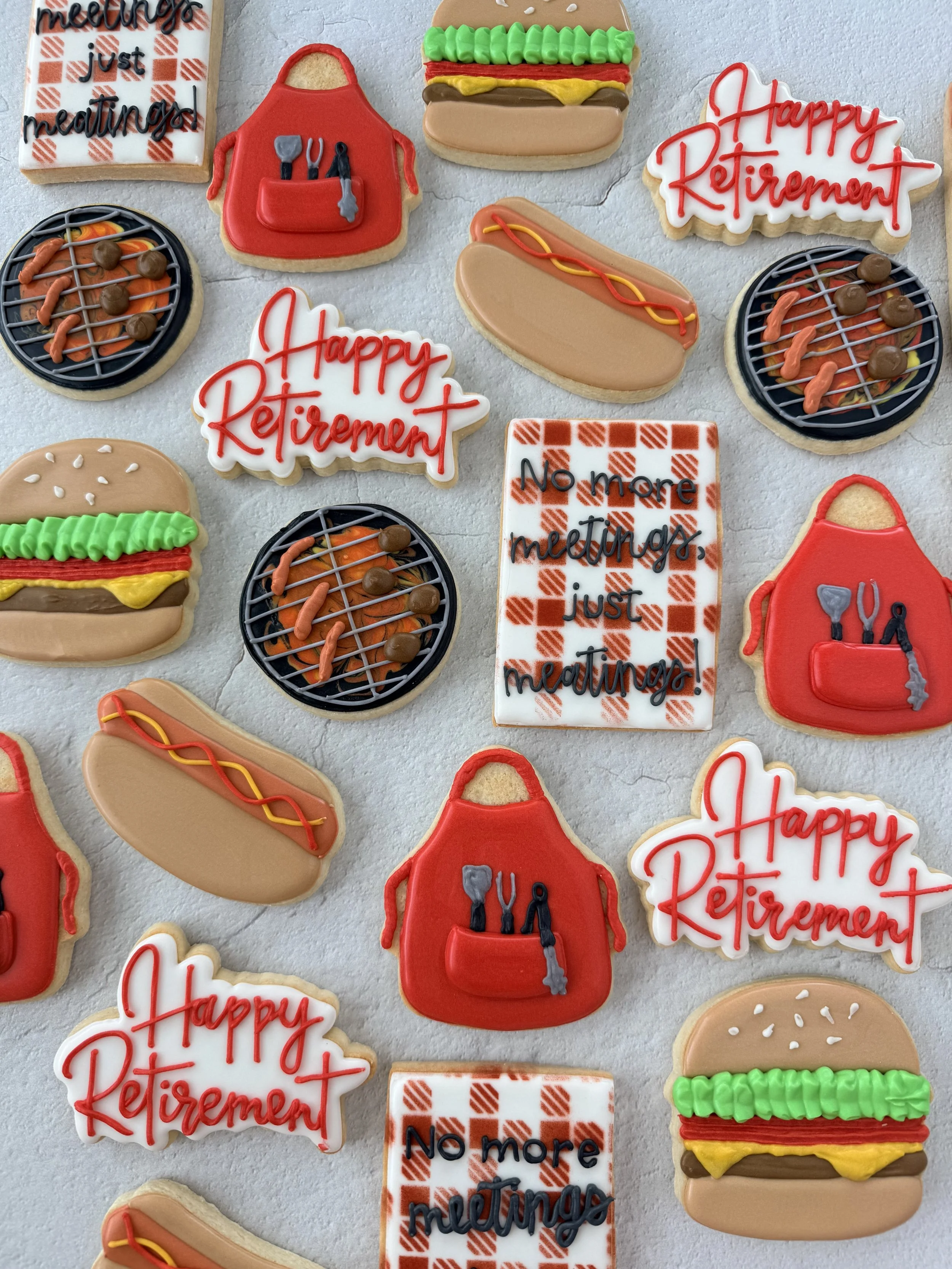 Decorative cookies in various shapes and designs, including hot dogs, hot dog buns, BBQ grills, a chef's apron with utensils, and signs with messages like 'Happy Retirement' and 'No more meetings'.