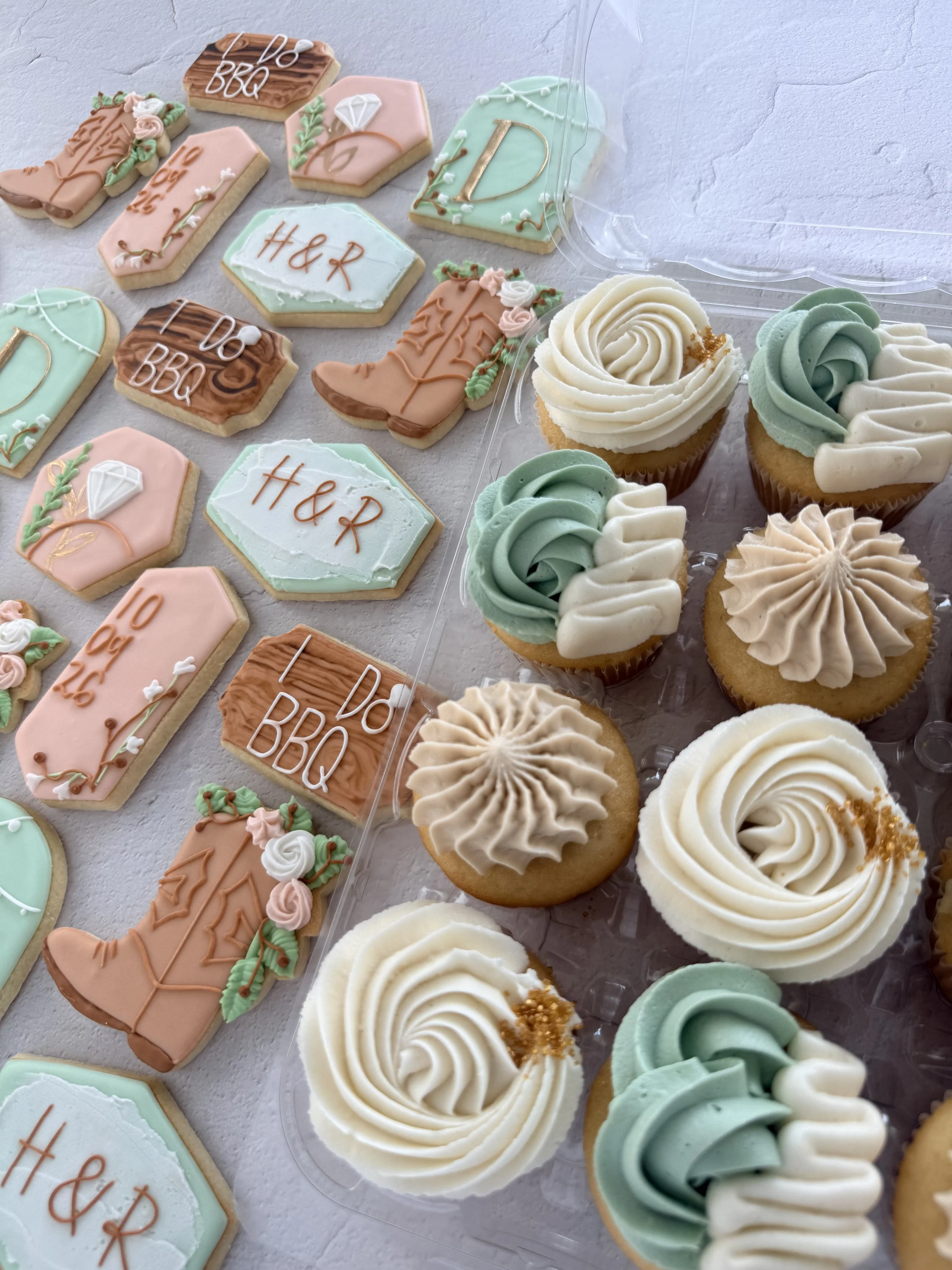 Assorted decorated cookies and cupcakes with wedding or engagement themes, featuring pastel colors, floral decor, and inscriptions like 'H&R' and 'I Do BBQ' on cookies, arranged on a light-colored surface.