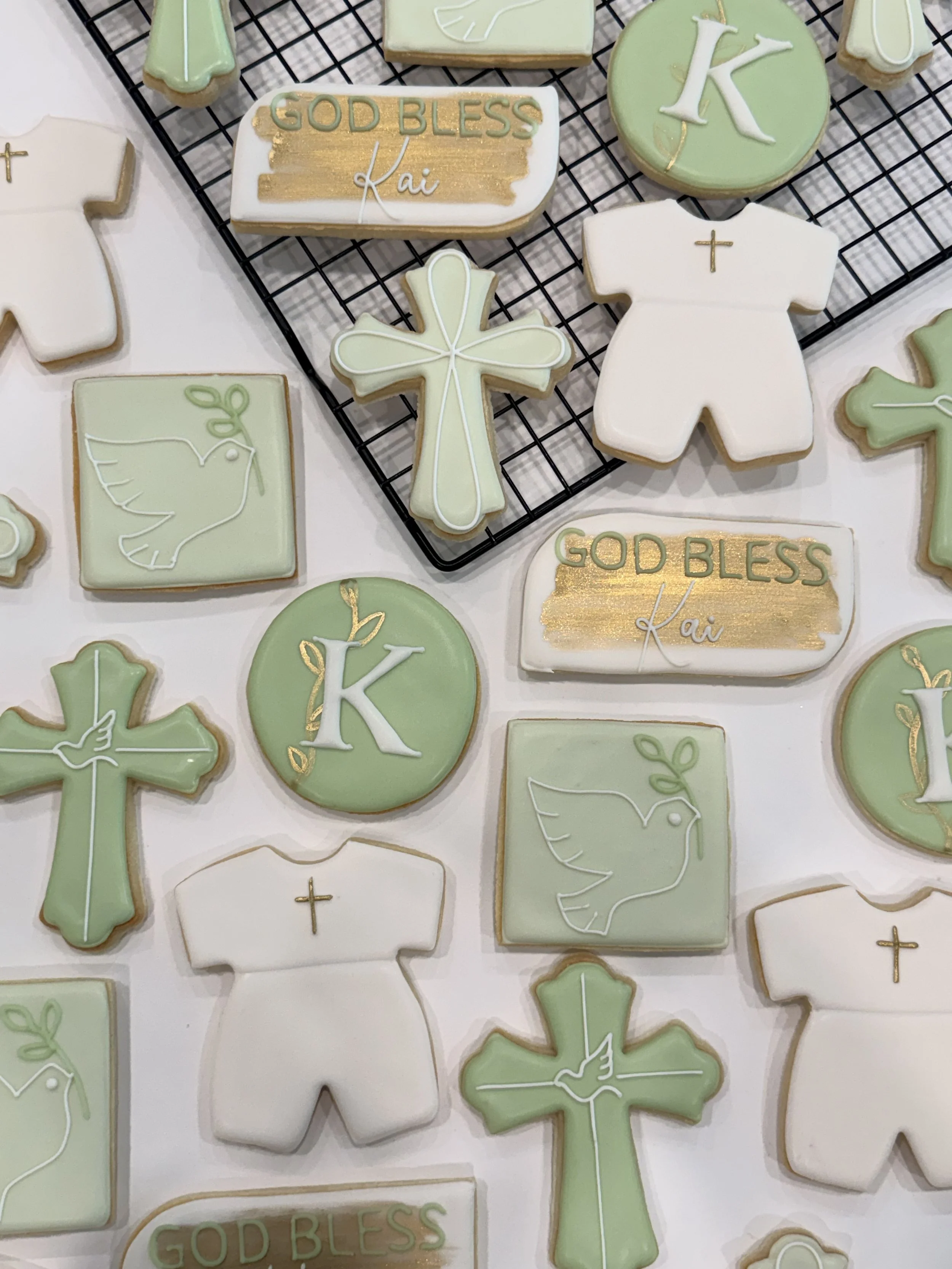 Decorated cookies in Christian symbols and messages placed on a cooling rack, including crosses, a t-shirt, a dove, a circle with the letter K, and a rectangular cookie with "GOD BLESS Kai" written in gold and green icing.